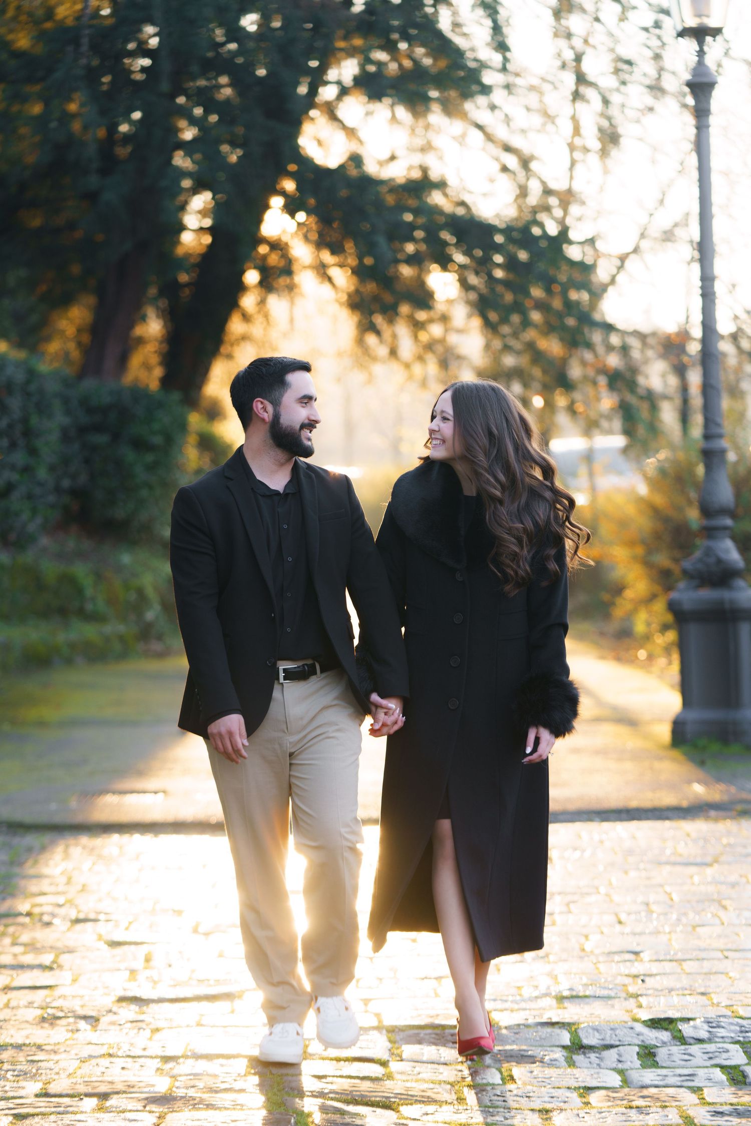 A couple walking together on a cobblestone path at sunset, holding hands and wearing stylish black coats.