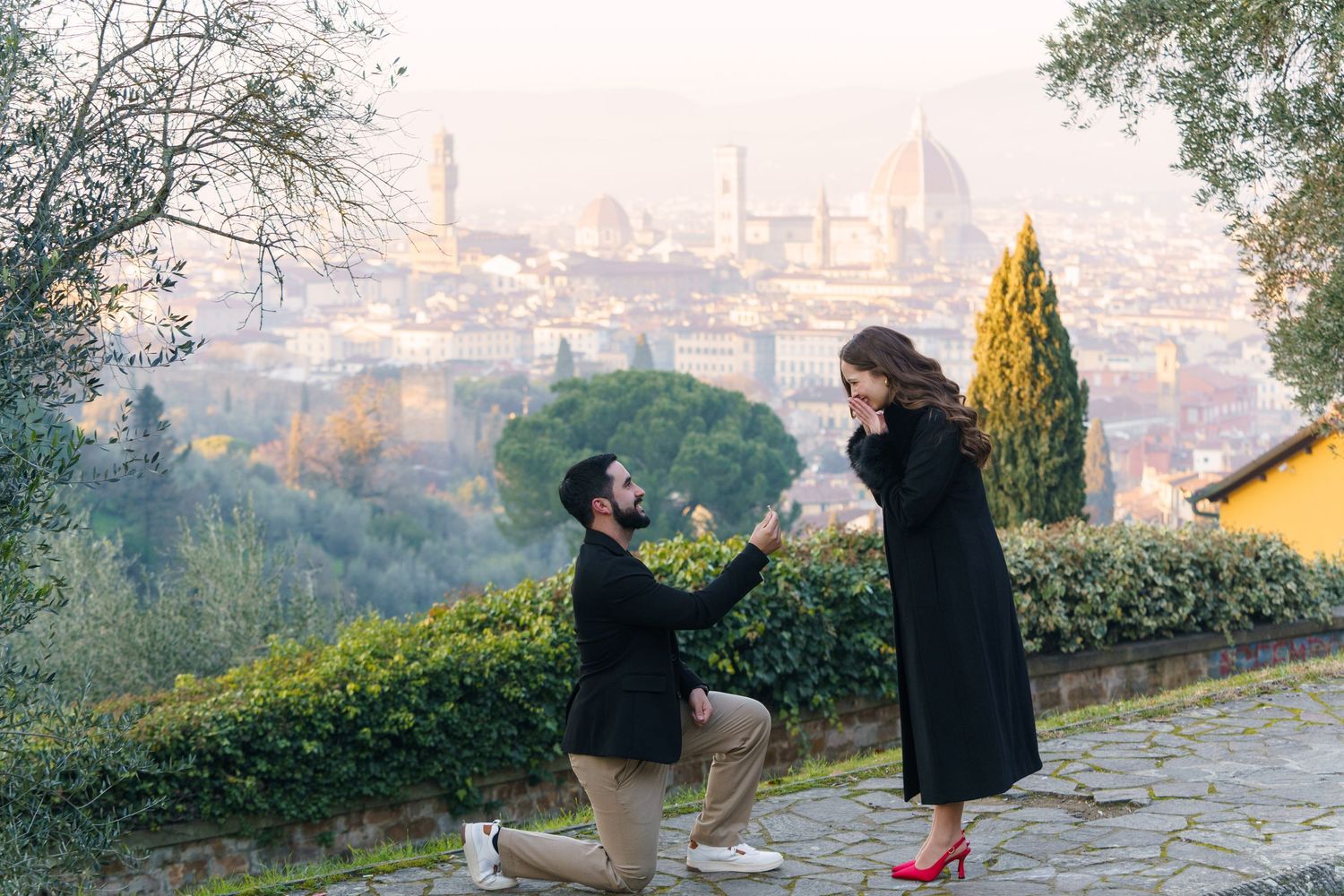 Two silhouettes enjoy sunset views over a misty European cityscape from an elevated viewpoint in a park.