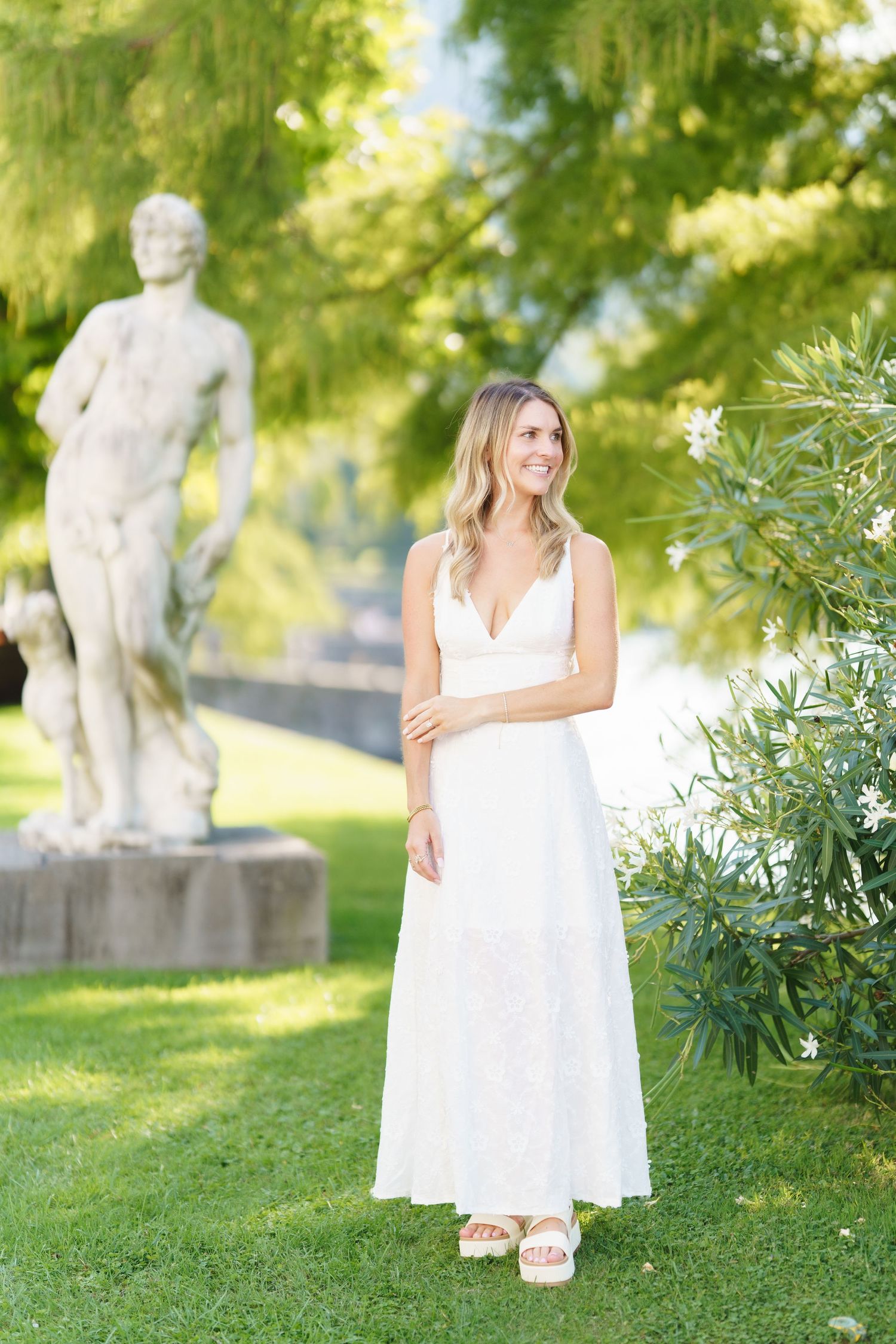 A figure in a flowing white dress poses gracefully in a sunlit garden with stone statues and lush greenery.