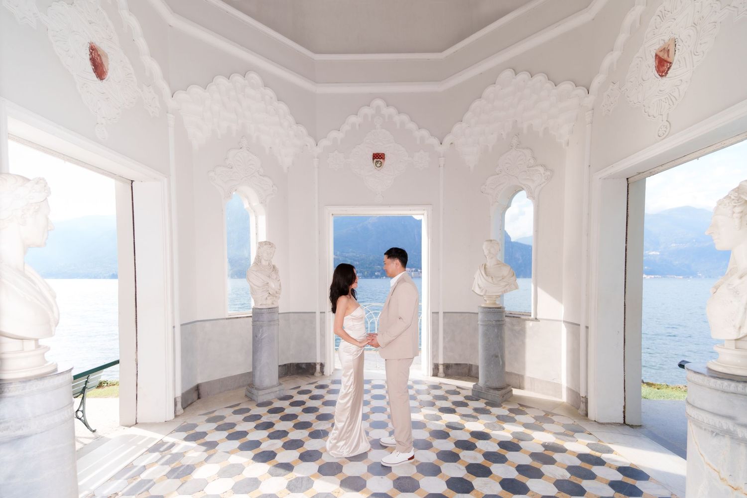 Two people in white outfits stand in an elegant white gazebo with a checkered floor overlooking a scenic ocean view.