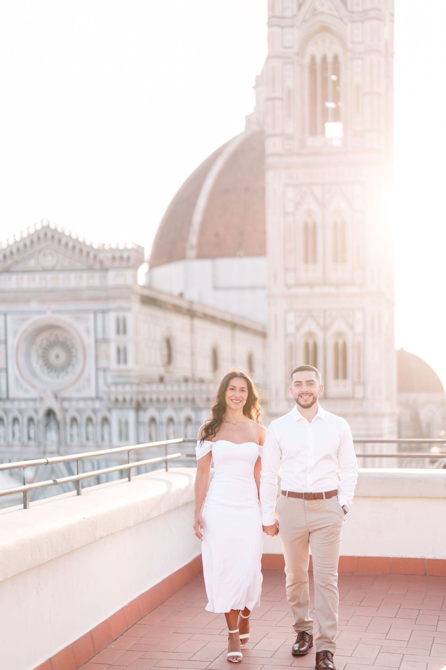 A couple in white outfits walks together on a rooftop terrace with a historic church spire visible in the background.