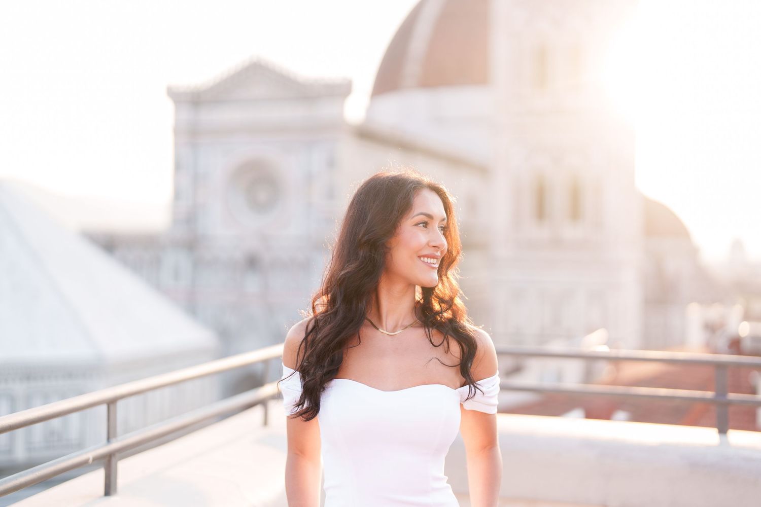 A woman in a white dress laughs joyfully while standing on a rooftop terrace during golden hour.