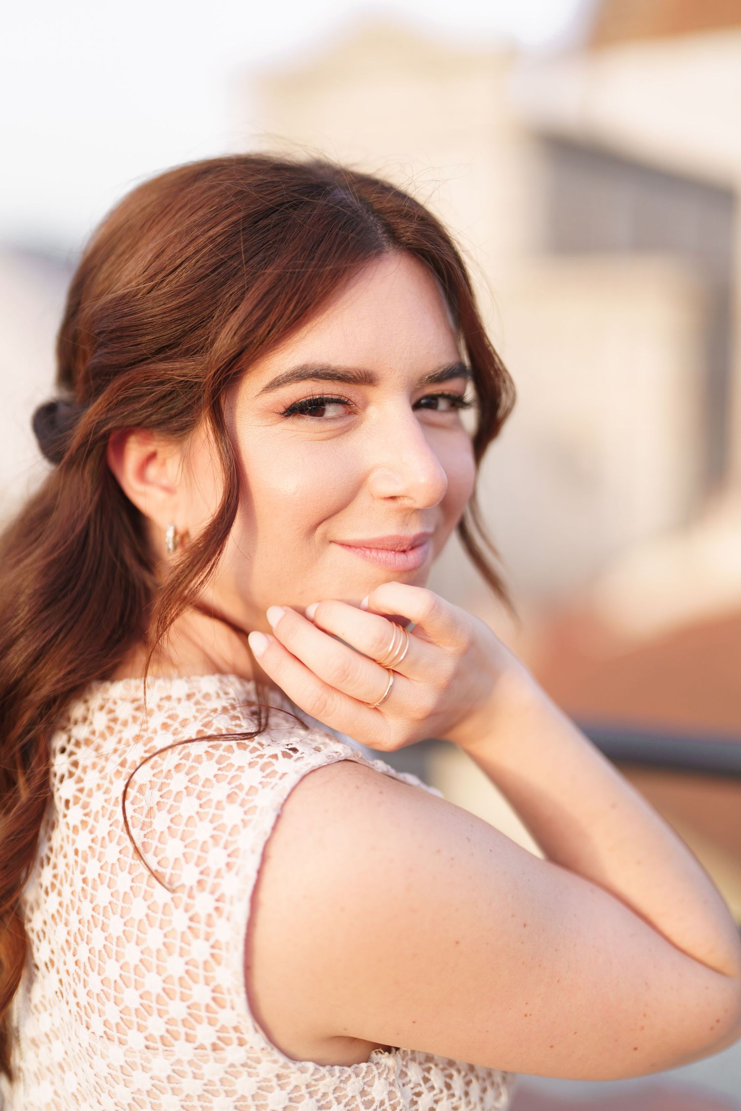 A person wearing a white lace sleeveless top smiles warmly while touching their face in soft sunlight.