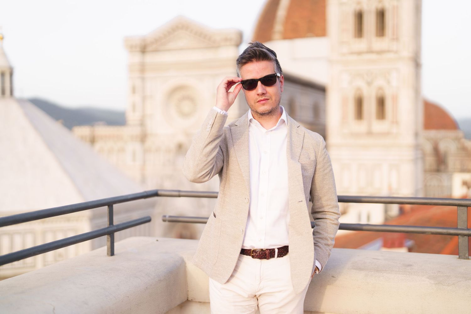 A person in a light colored suit stands on a balcony overlooking a European city with historic architecture.