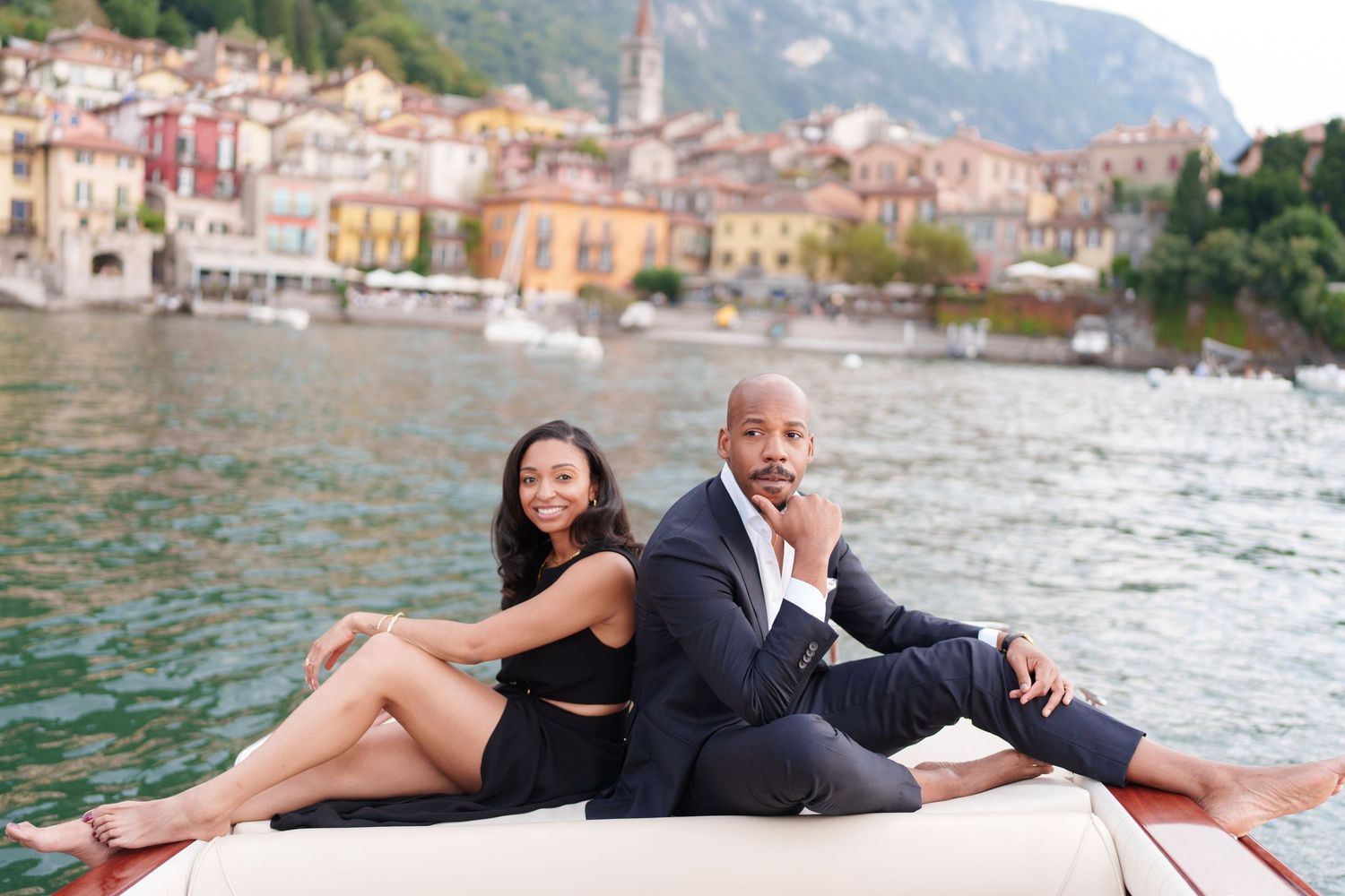 A couple in formal attire poses romantically on a boat dock by a scenic waterfront with Italian buildings in the background.