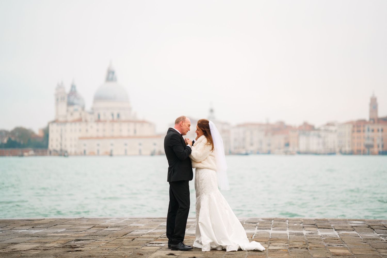 A couple shares a romantic moment along the Venice waterfront with the iconic cityscape and lagoon in the background.