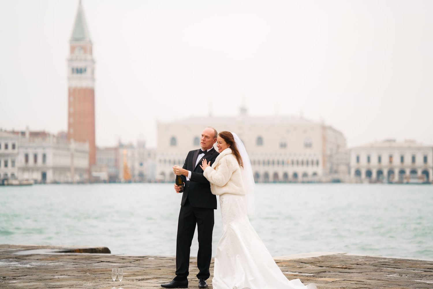 A romantic couple in formal wedding attire embraces while standing on a pier overlooking Venice's iconic waterfront skyline.
