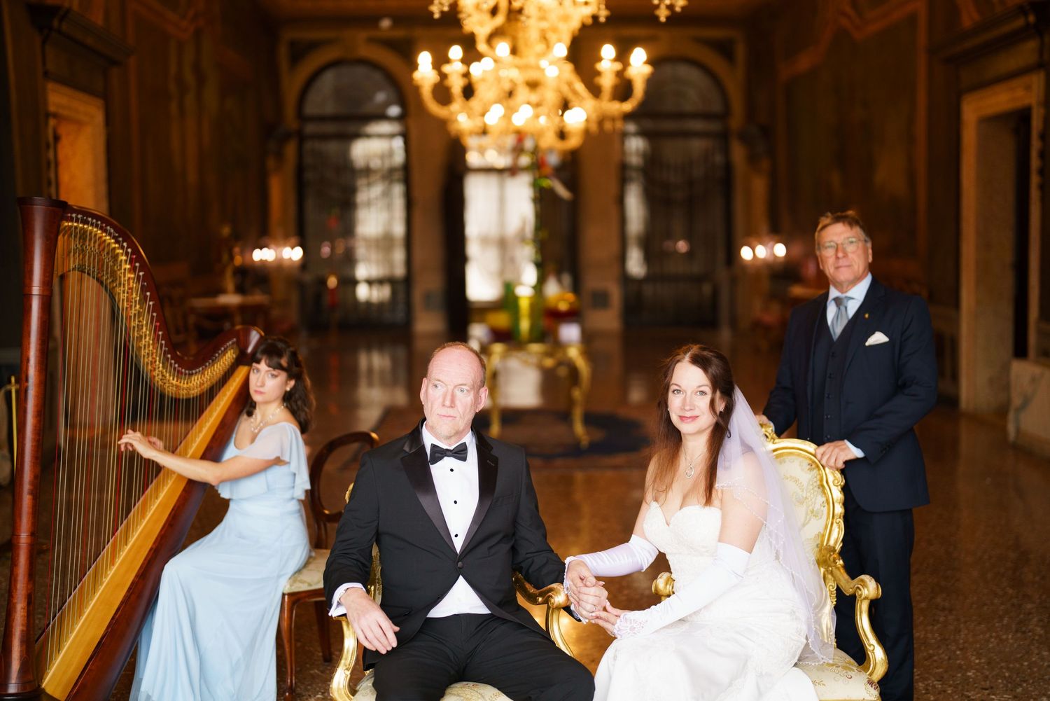 A couple shares their first dance in an elegant ballroom with a chandelier overhead and a harpist playing in the background.