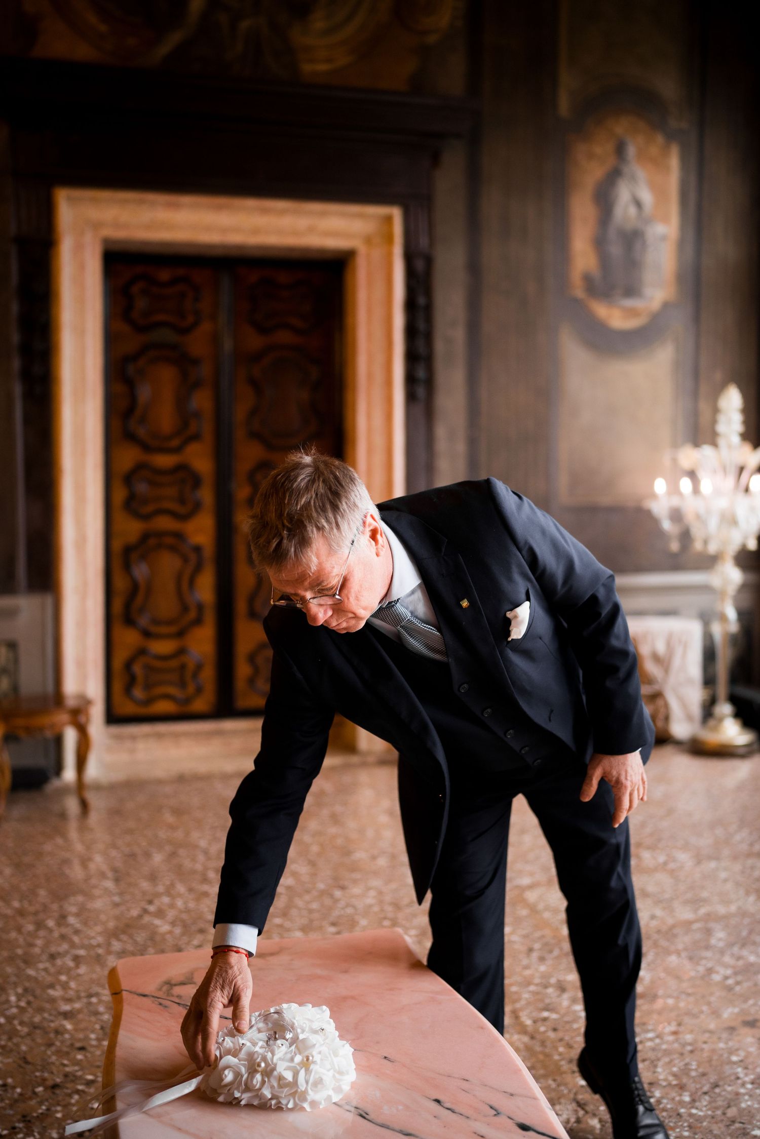 A person in a dark suit bends down to inspect a white wedding cake on a carpeted floor in an elegant room.