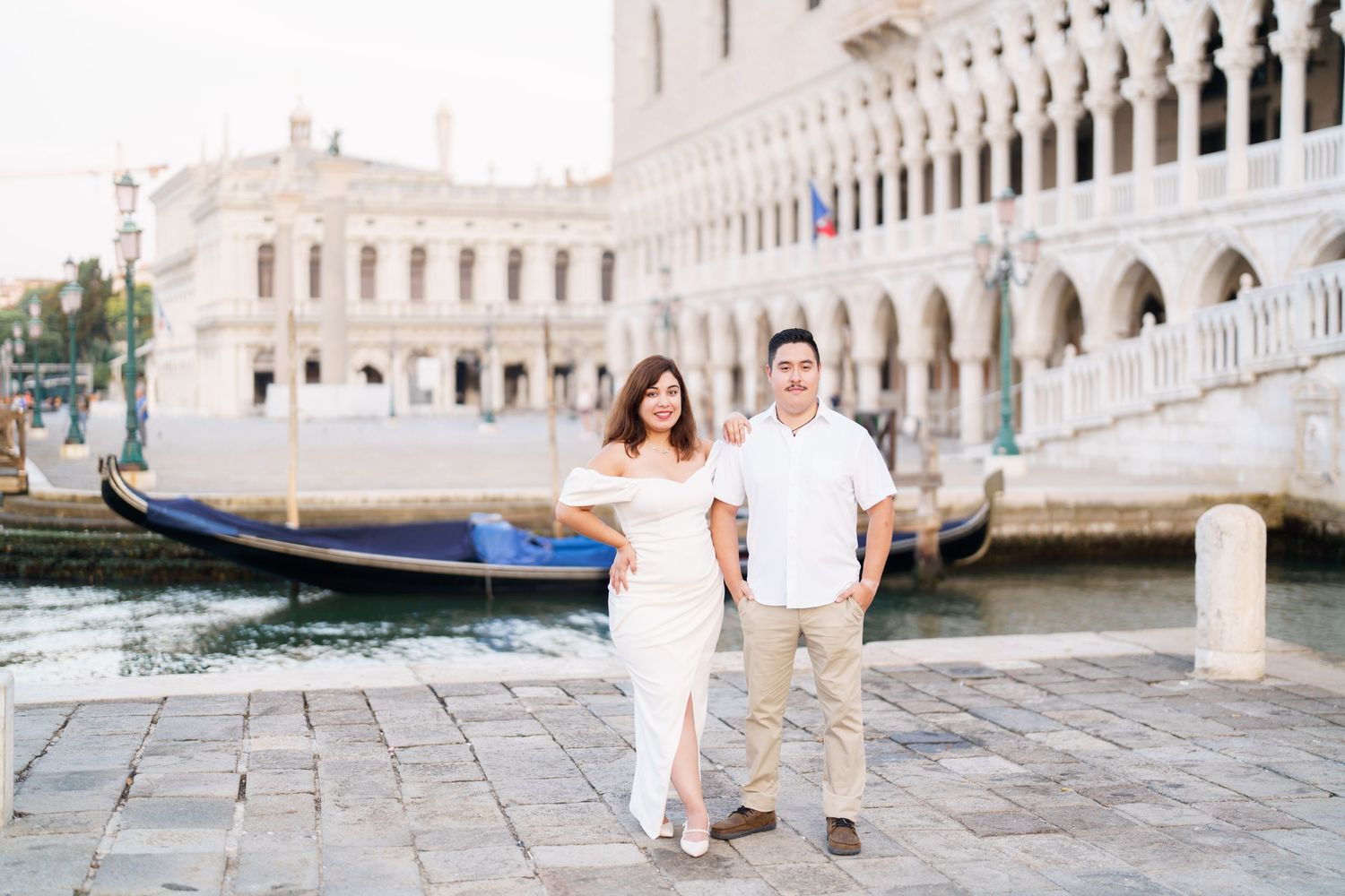 A couple in white outfits poses together on a historic bridge in Venice with gondolas and architecture in the background.