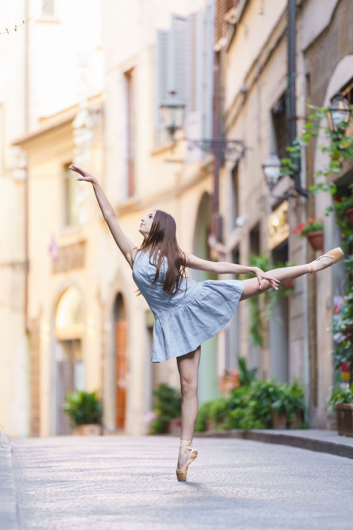 photographer Florence A ballet dancer in a blue dress performs an arabesque on pointe in a charming European street setting.