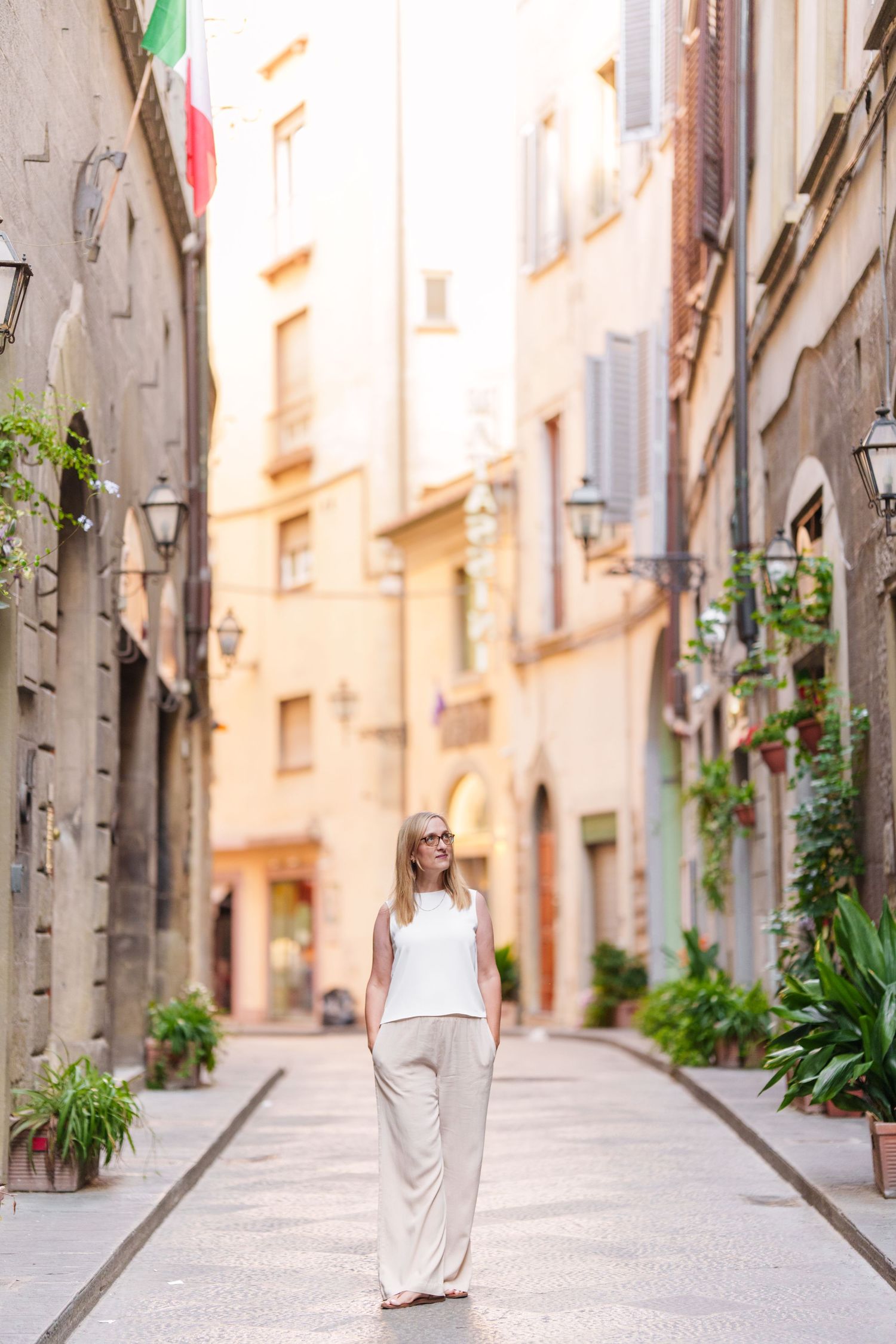 Woman in white dress walks through charming narrow European street with stone walls and potted plants.