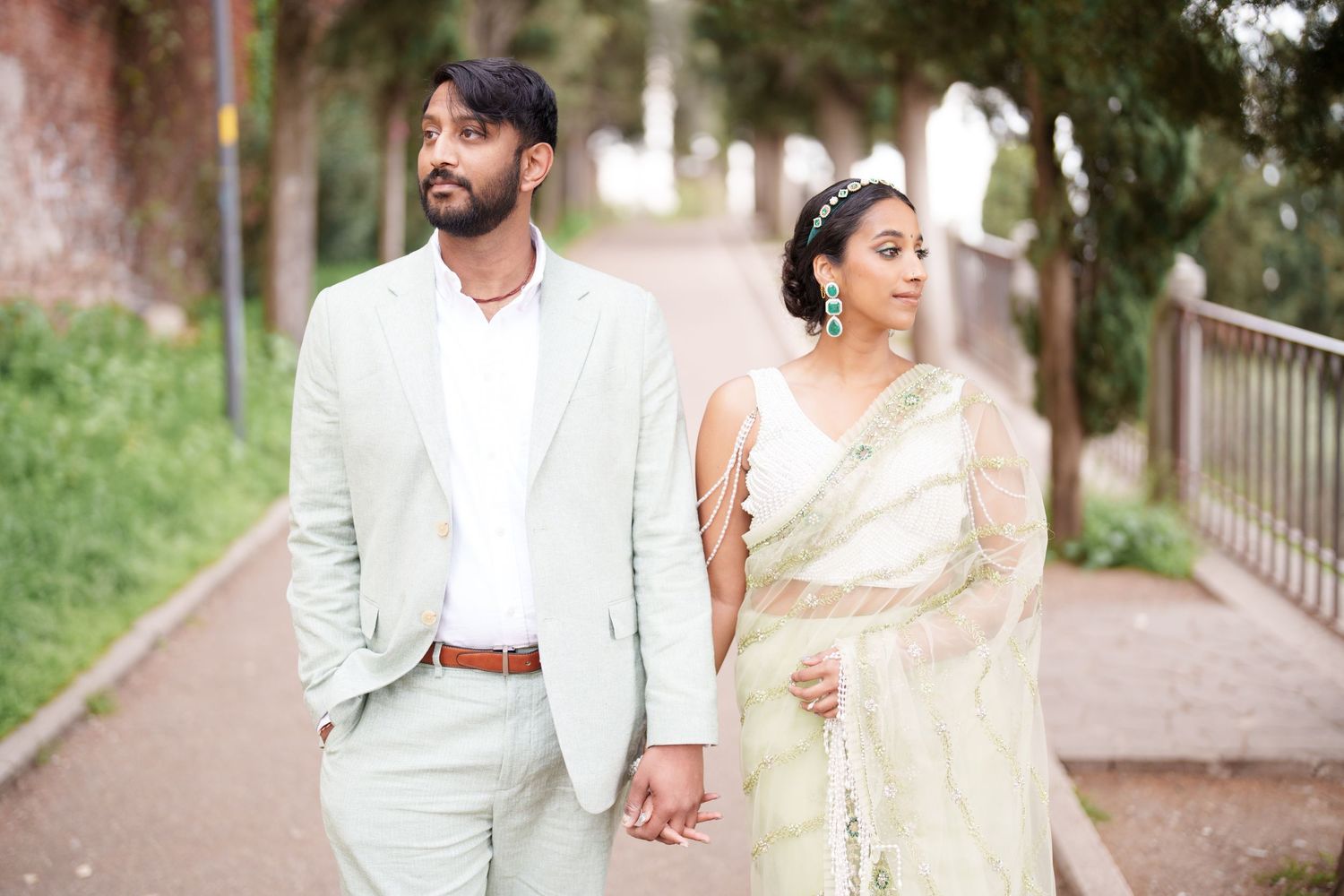 photographer verona A couple in elegant white and cream outfits walk together on a tree-lined path during a romantic photoshoot.