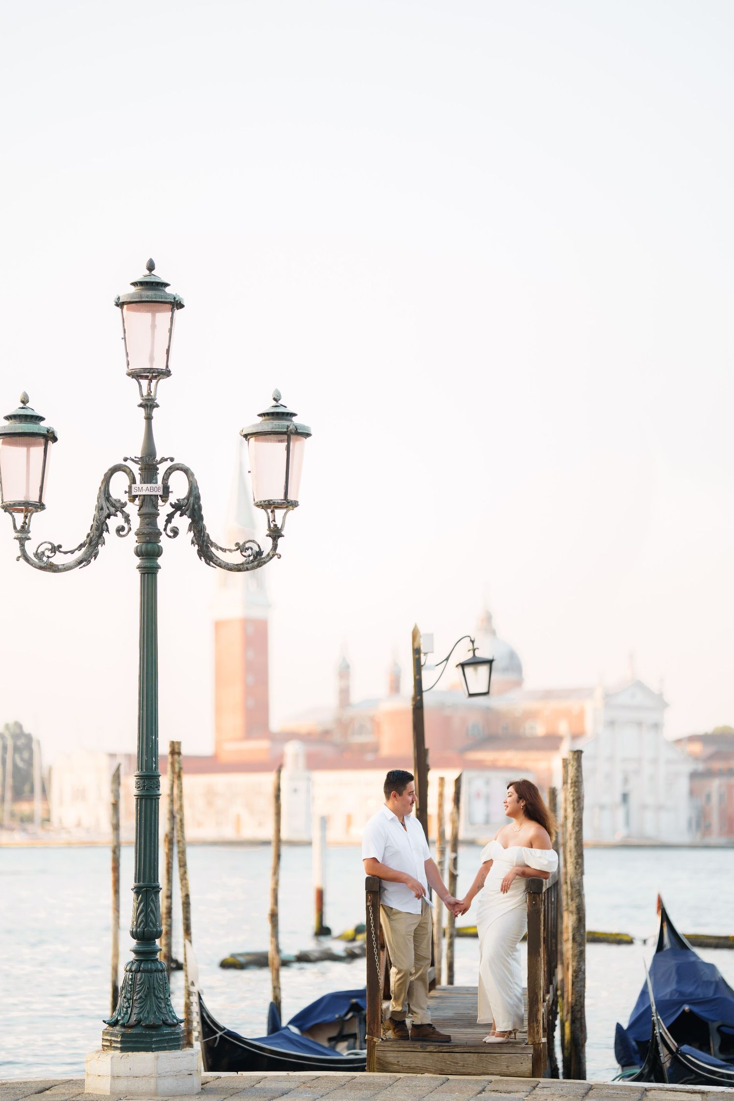 Romantic couple stands on a Venetian dock with ornate street lamp and gondolas against the city skyline at sunset.