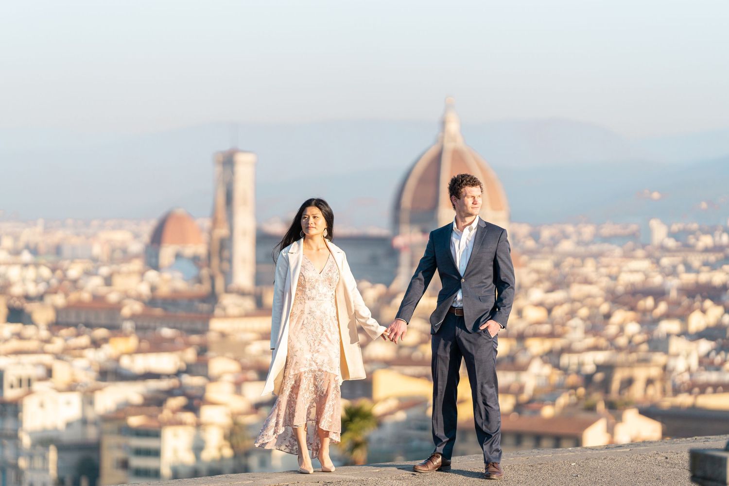 A couple holds hands at a scenic viewpoint overlooking Florence, Italy with the Duomo cathedral visible in the background.