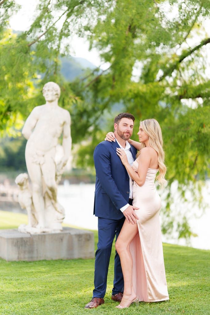A couple poses romantically on a grassy lawn with white statues and willow trees in the background.