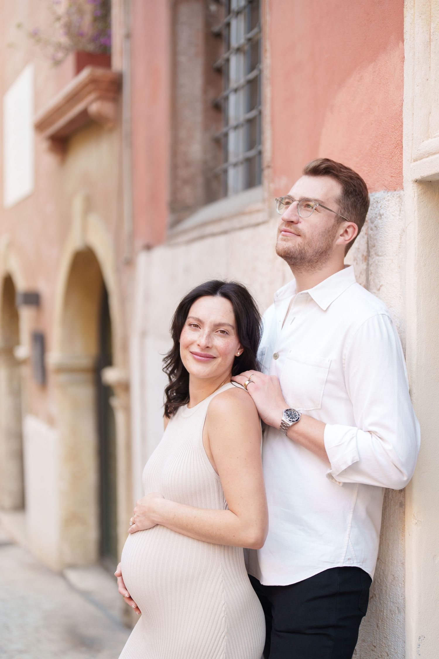 Romantic couple embraces against pink stone wall in European alleyway during engagement photo session.