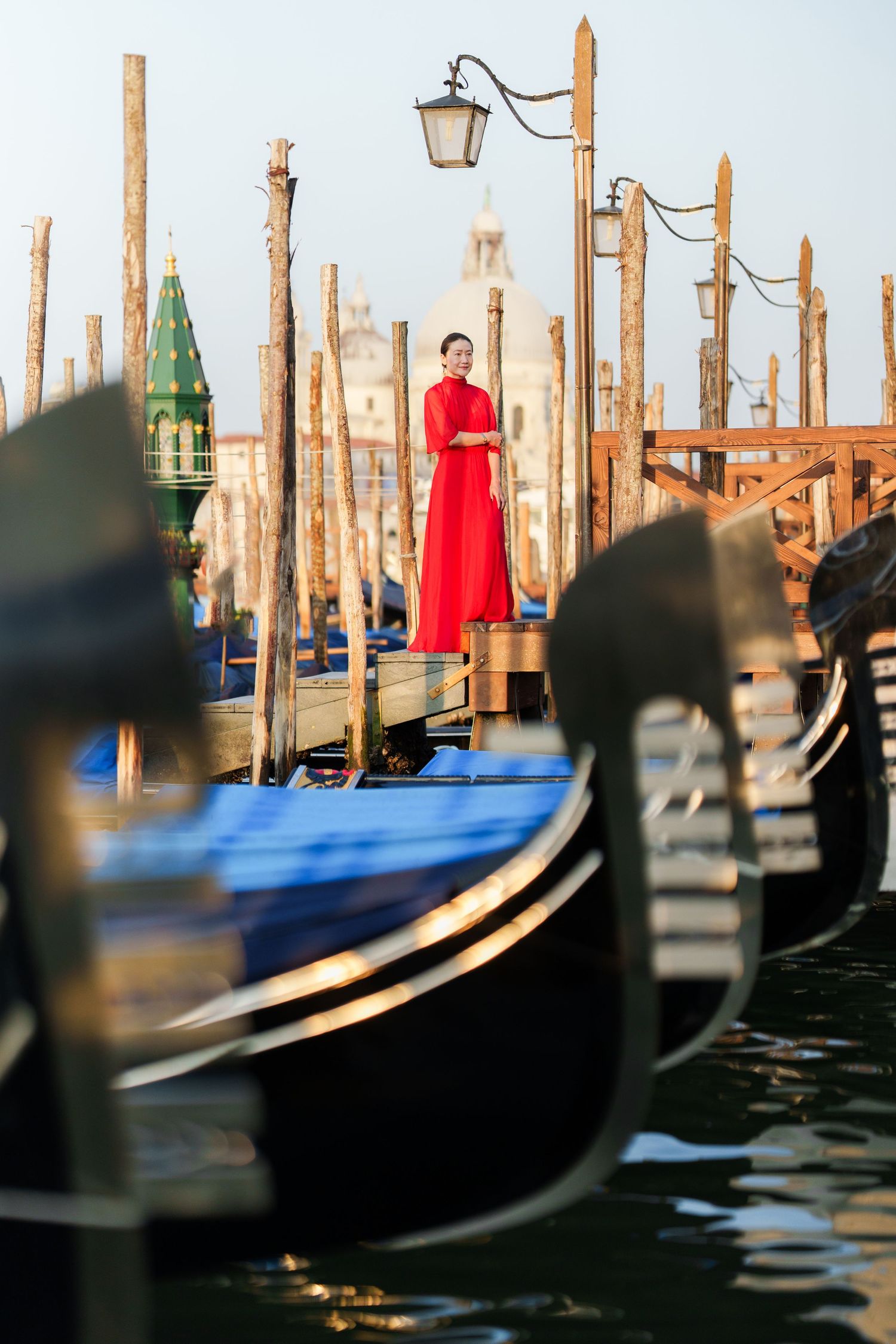 Woman in red dress standing on Venice dock surrounded by gondolas with San Giorgio Maggiore church in background.