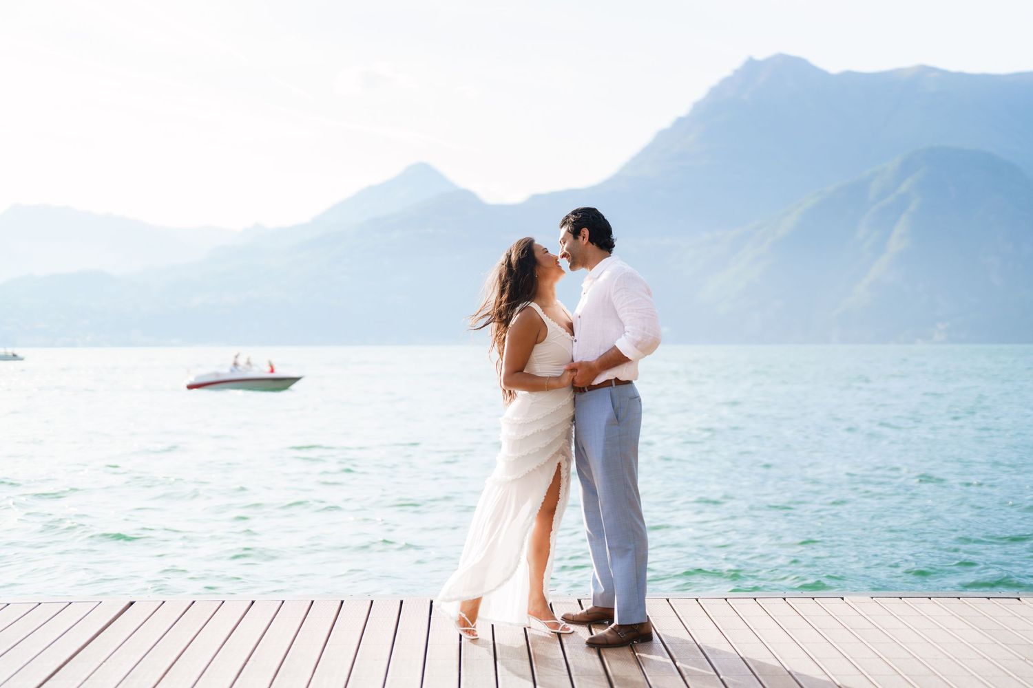 Couple kissing on a lakeside dock with mountains and turquoise water in the background.
