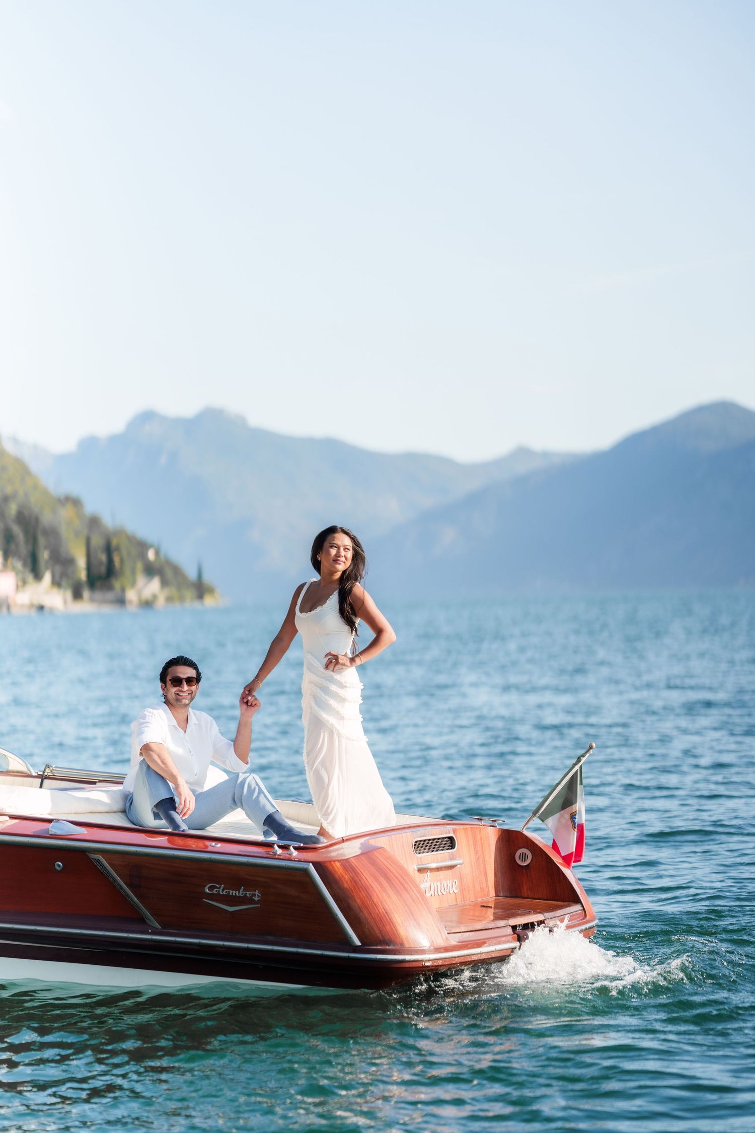 Couple in white attire on a classic wooden speedboat on a serene mountain lake on a sunny day.