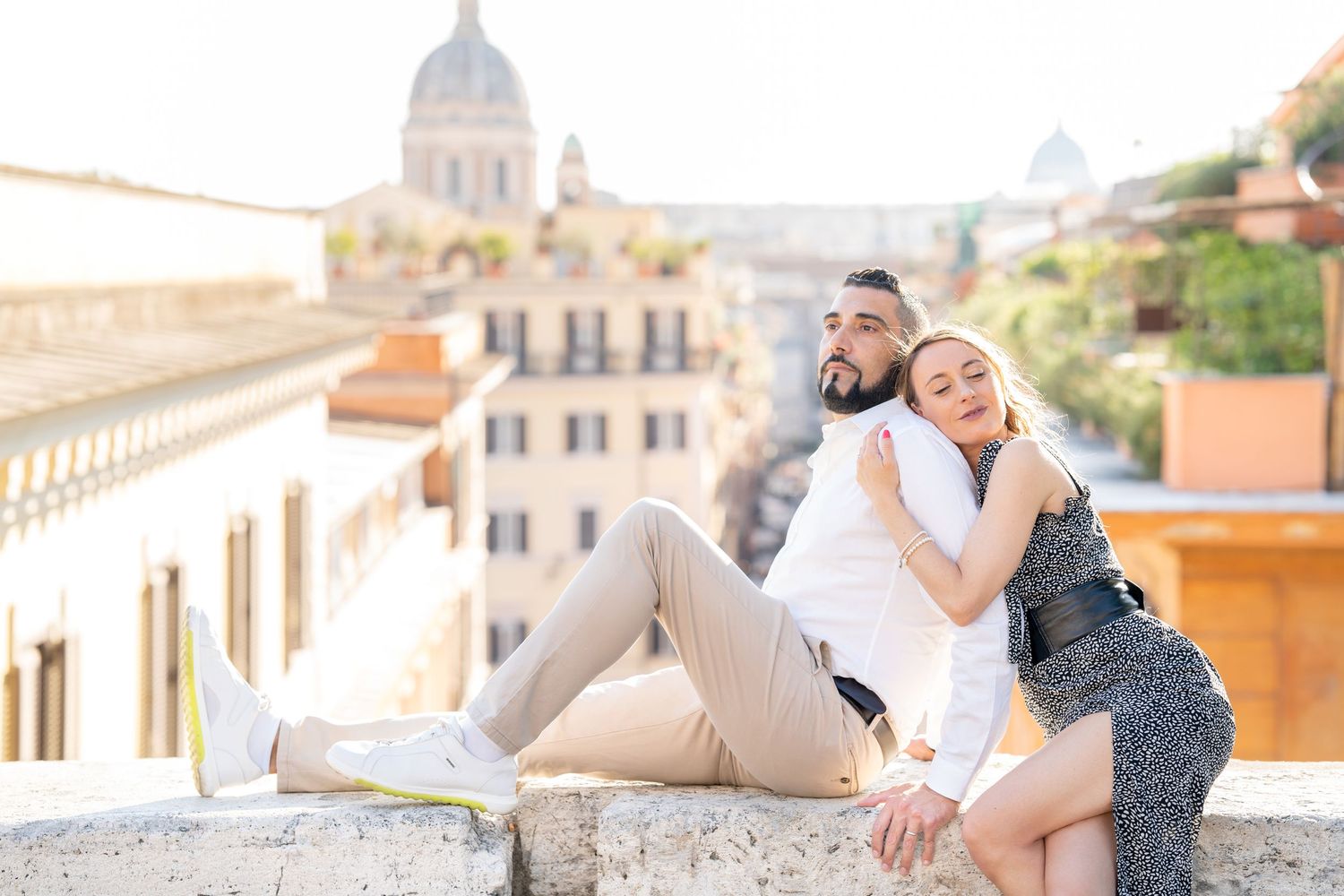 Couple sitting on a stone wall in Rome with historic buildings and a dome in the background.