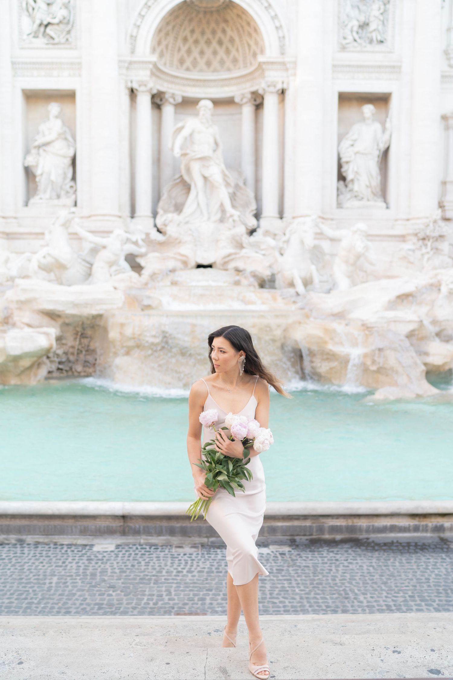 Woman in white dress holding pink peonies at the Trevi Fountain in Rome, Italy.