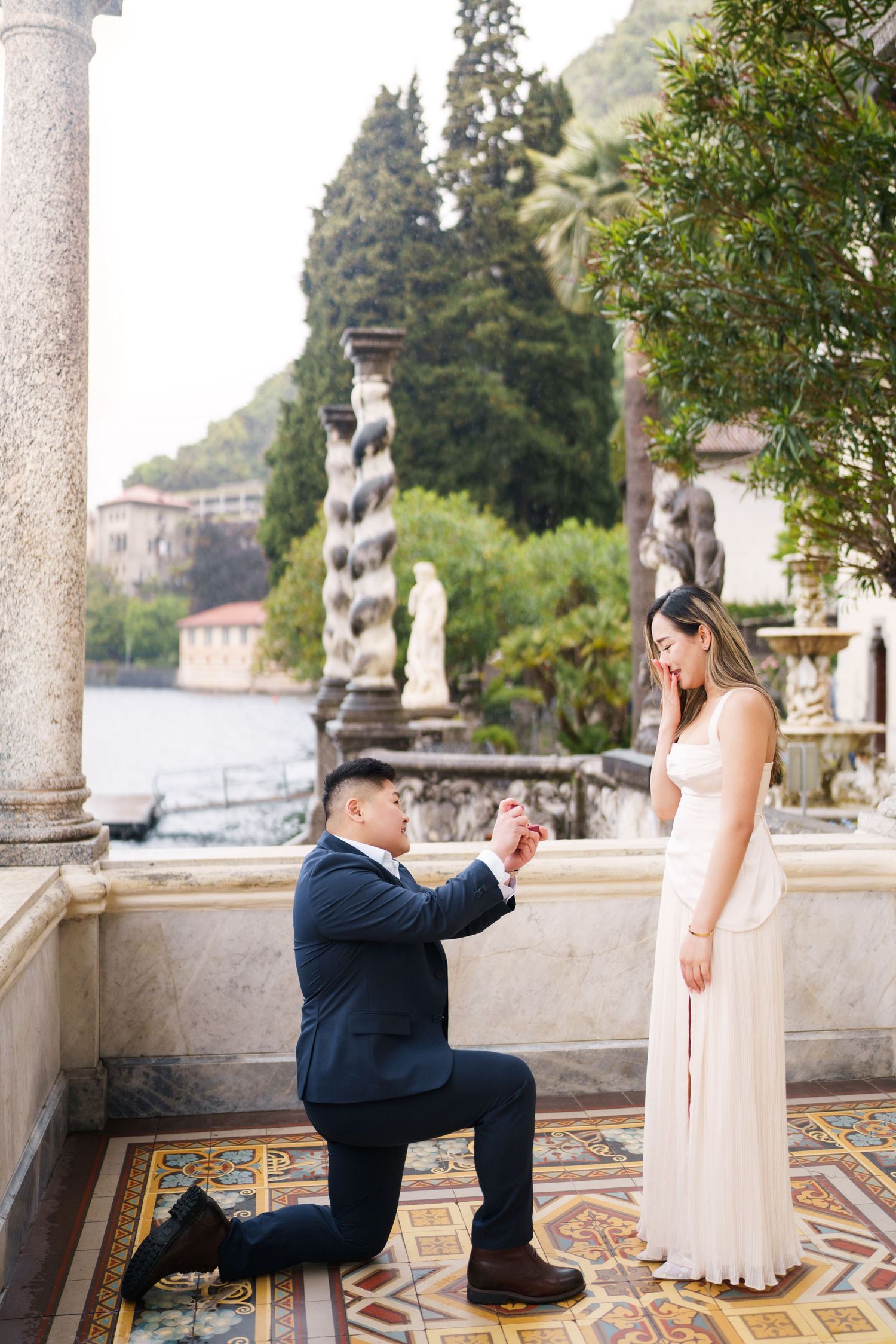 Man proposes on one knee to woman in elegant dress on ornate terrace overlooking Lake Como, Italy.