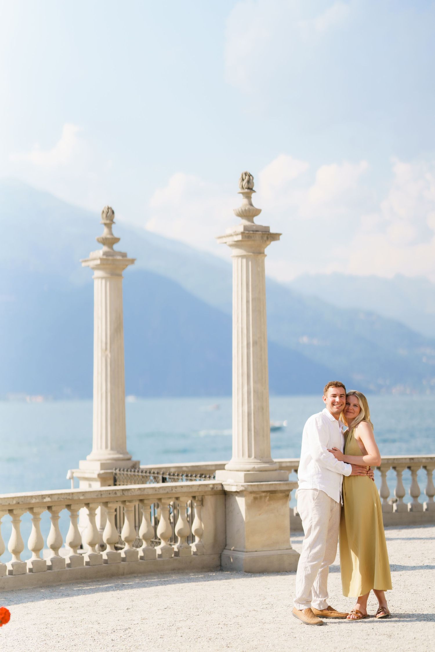 Couple embracing on elegant lakeside terrace with classical columns and mountain backdrop.