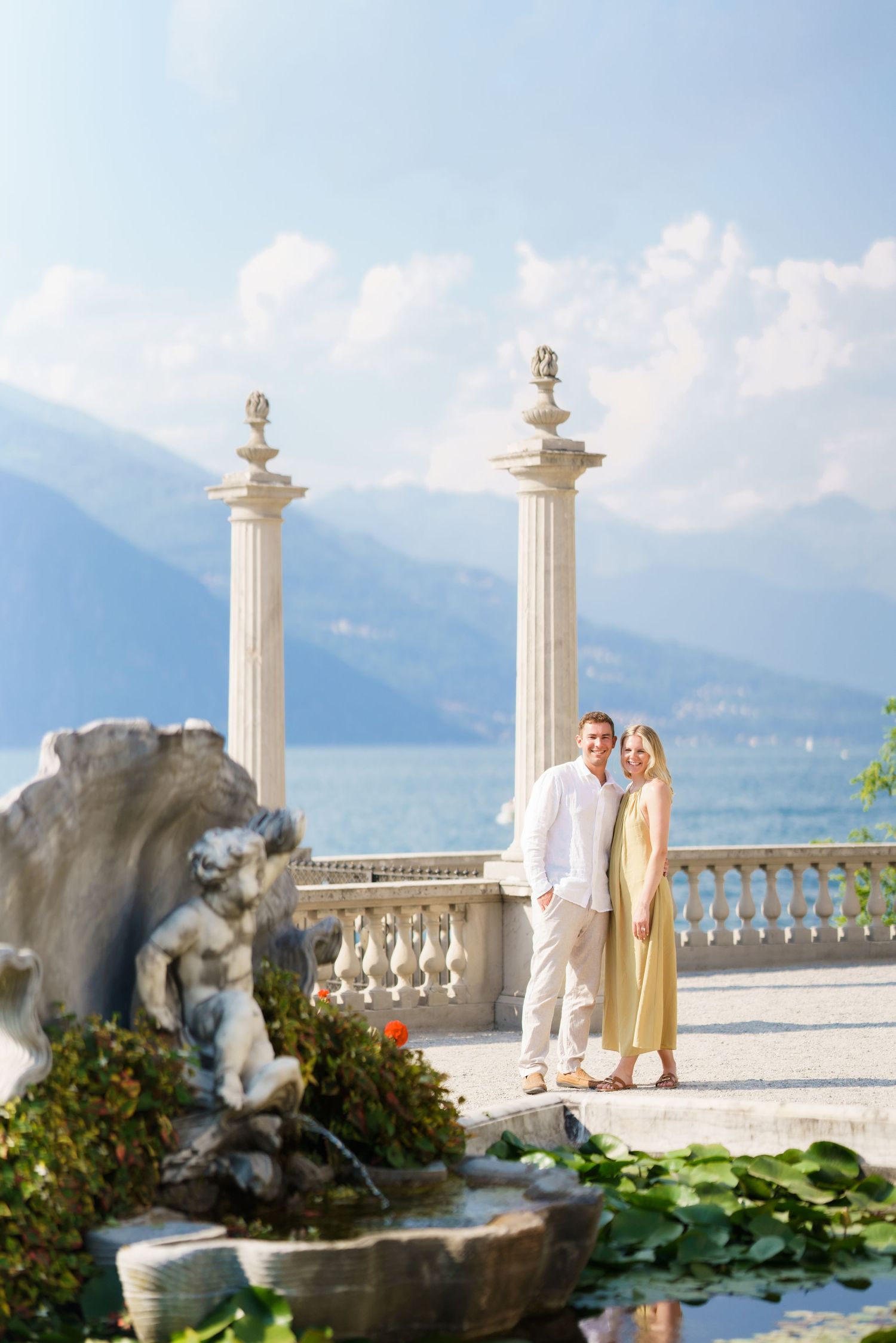 Couple posing on elegant Italian villa terrace with marble columns, cherub fountain, and scenic lake and mountain backdrop.