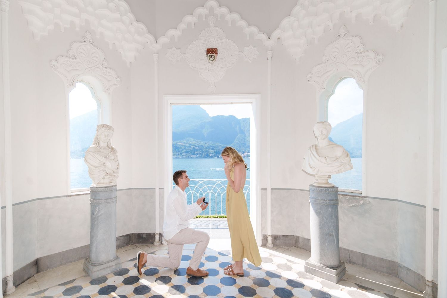 Man proposing to woman in elegant white villa with marble busts and stunning lake view backdrop.