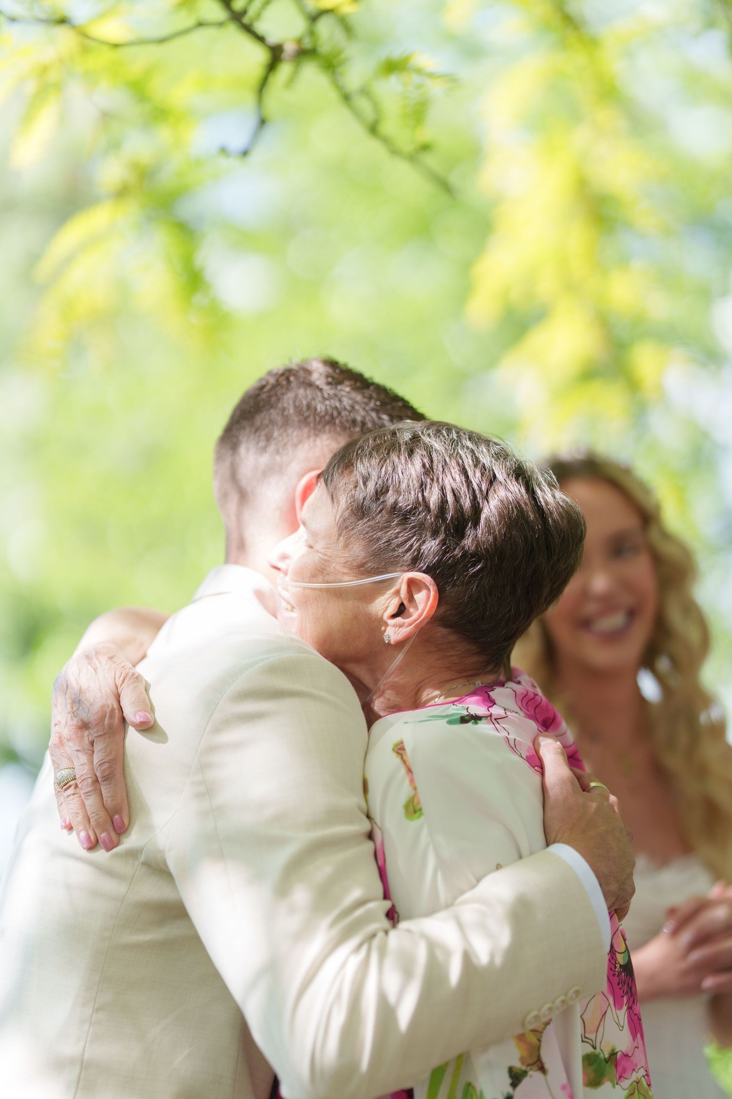 Groom in cream suit embracing older woman in floral dress at outdoor wedding, smiling bride visible in background.