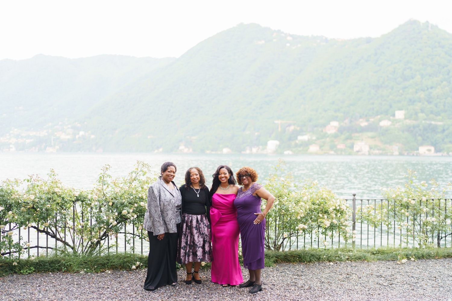 Four women posing together outdoors by a scenic lake with lush green hills and mountains in the background.