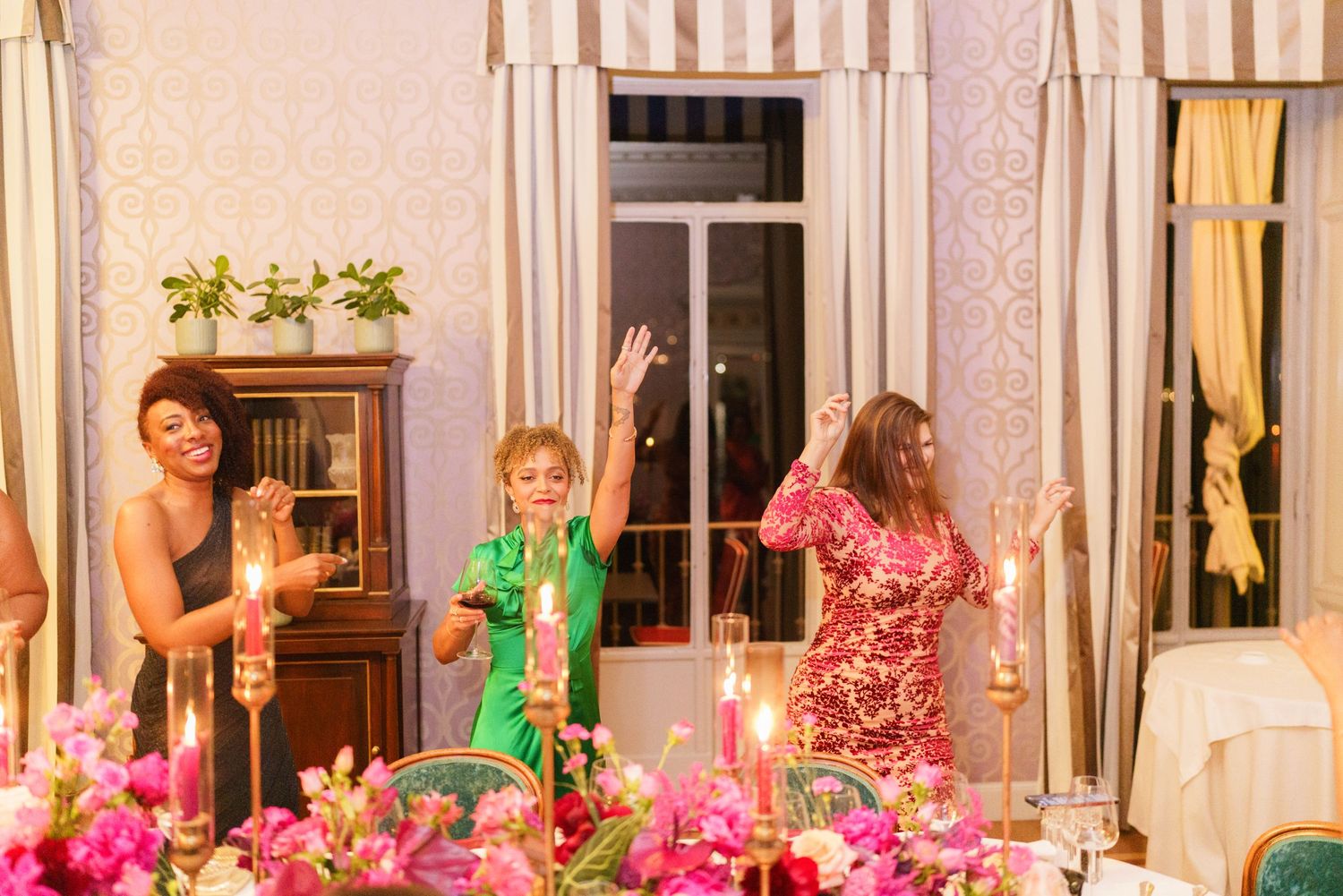 Three women celebrating at an elegant dinner party with candles and pink floral centerpieces.