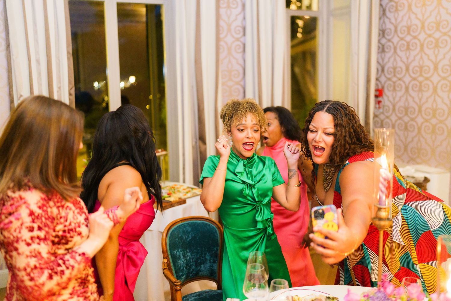 Women in colorful dresses laughing and celebrating together at an elegant indoor party event.