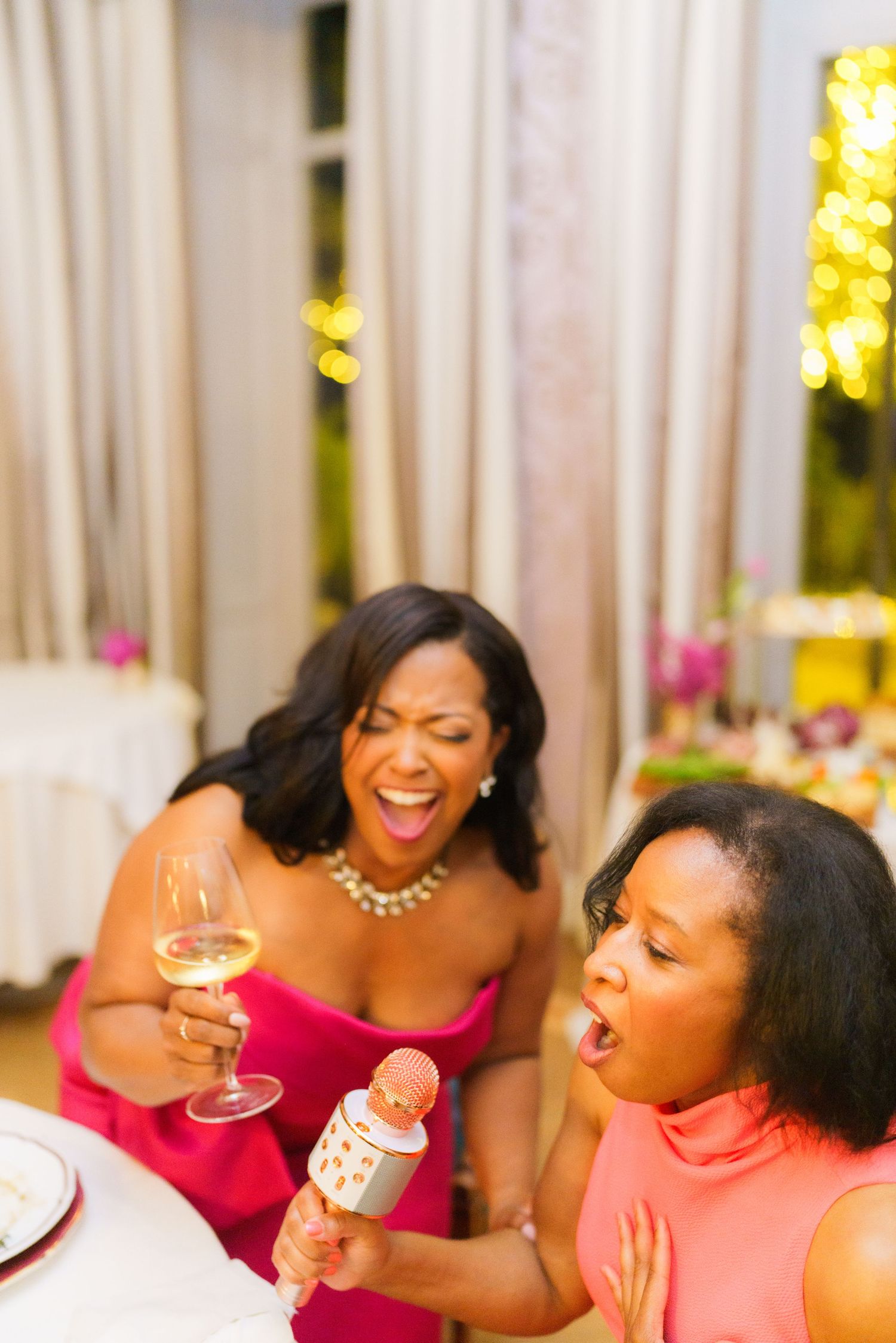 Two women laughing and singing karaoke at a party, one holding a wine glass and the other a microphone.