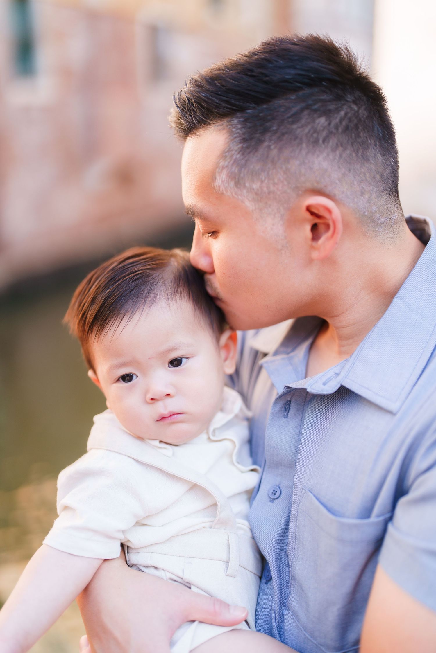 Asian father tenderly kissing his baby on the forehead while holding the infant outdoors near a canal.