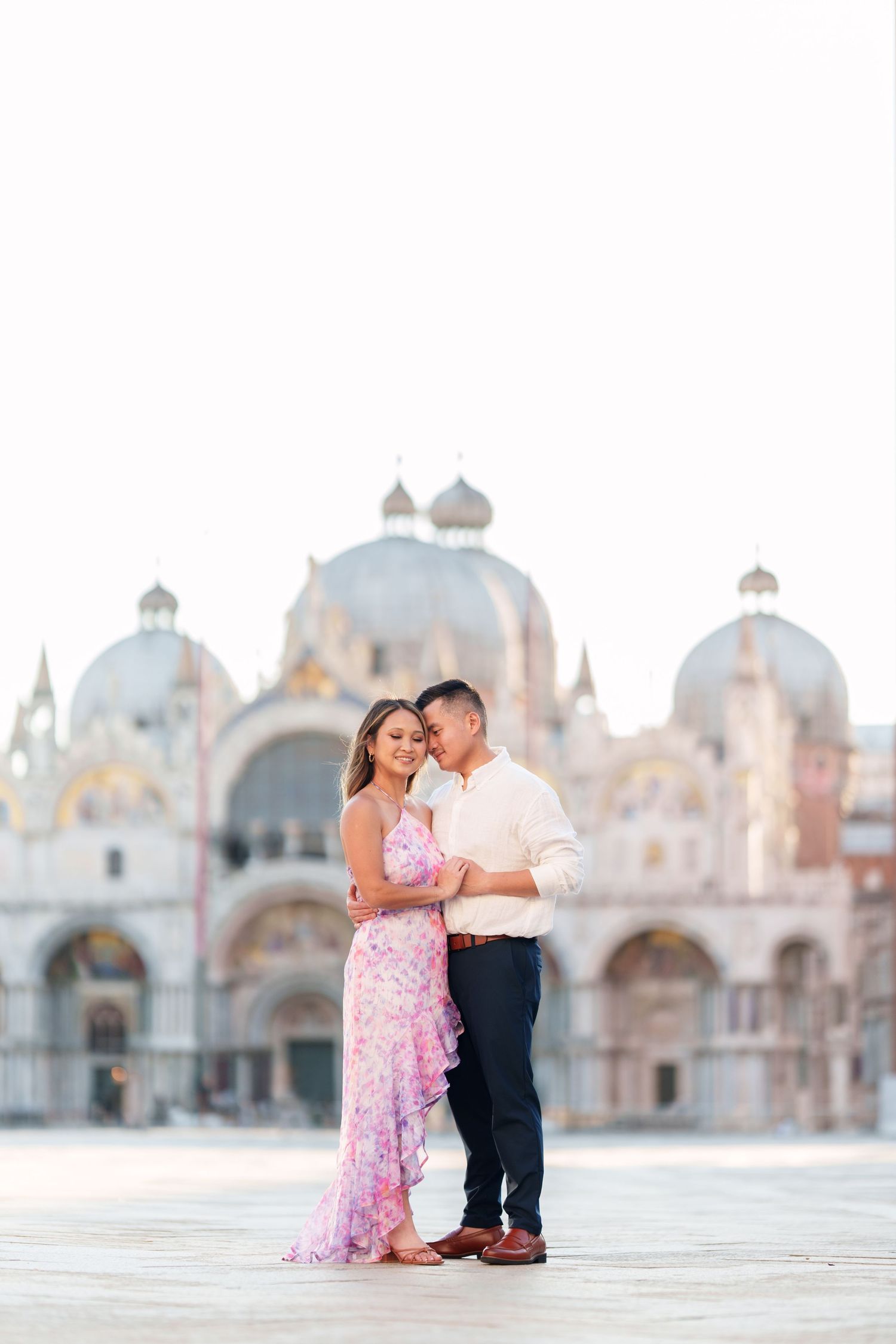 Couple embracing in St. Mark's Square, Venice, woman in pink floral dress, man in white shirt, Basilica backdrop.