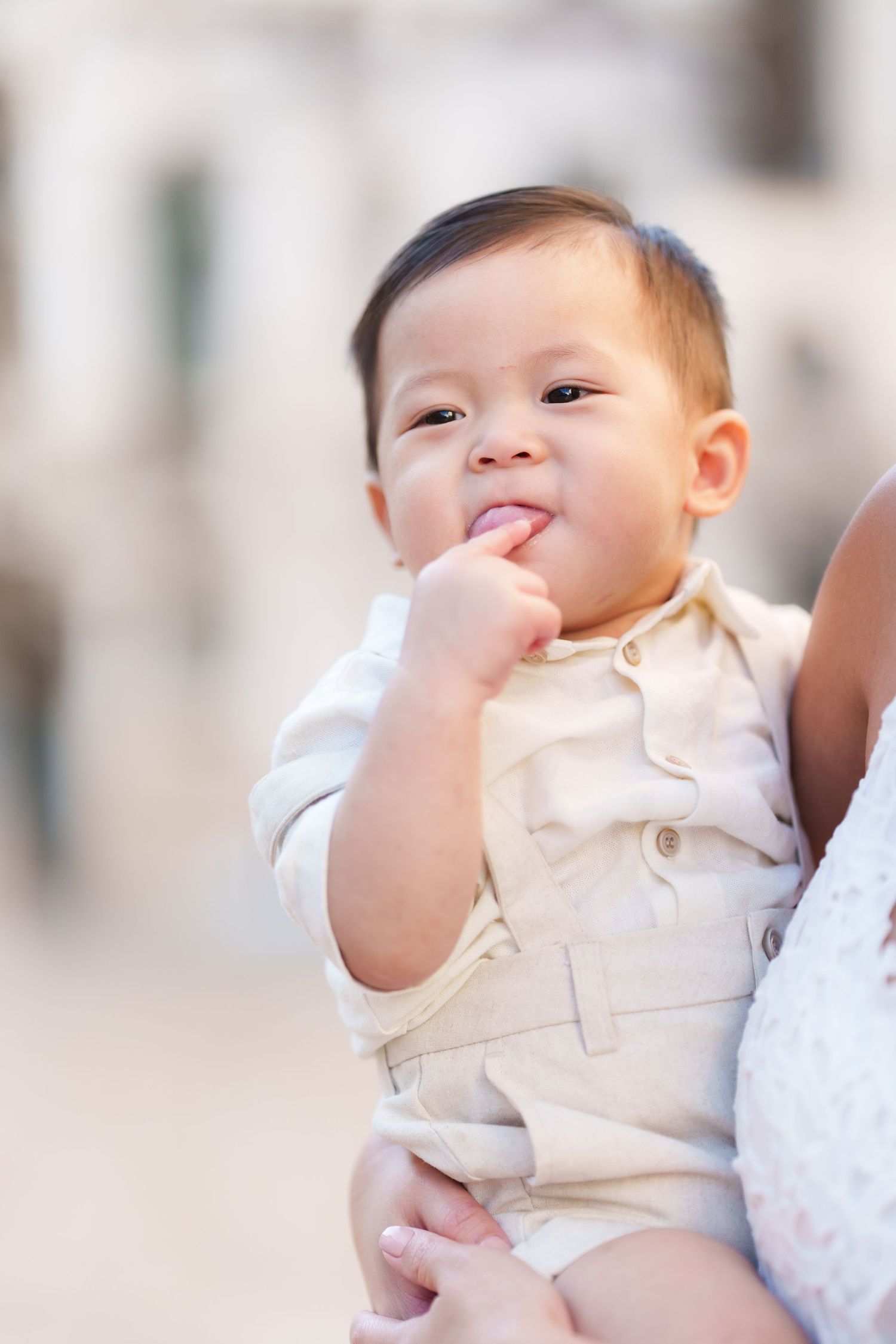 Adorable Asian baby in beige outfit smiling with finger in mouth, being held by an adult outdoors.