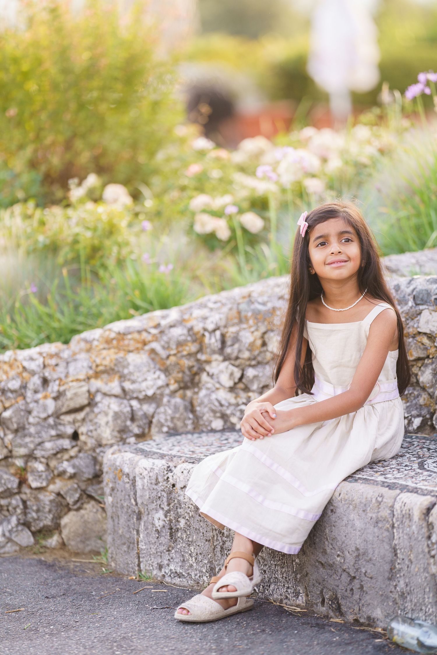 Young girl in white dress sitting on stone steps surrounded by blooming garden flowers.