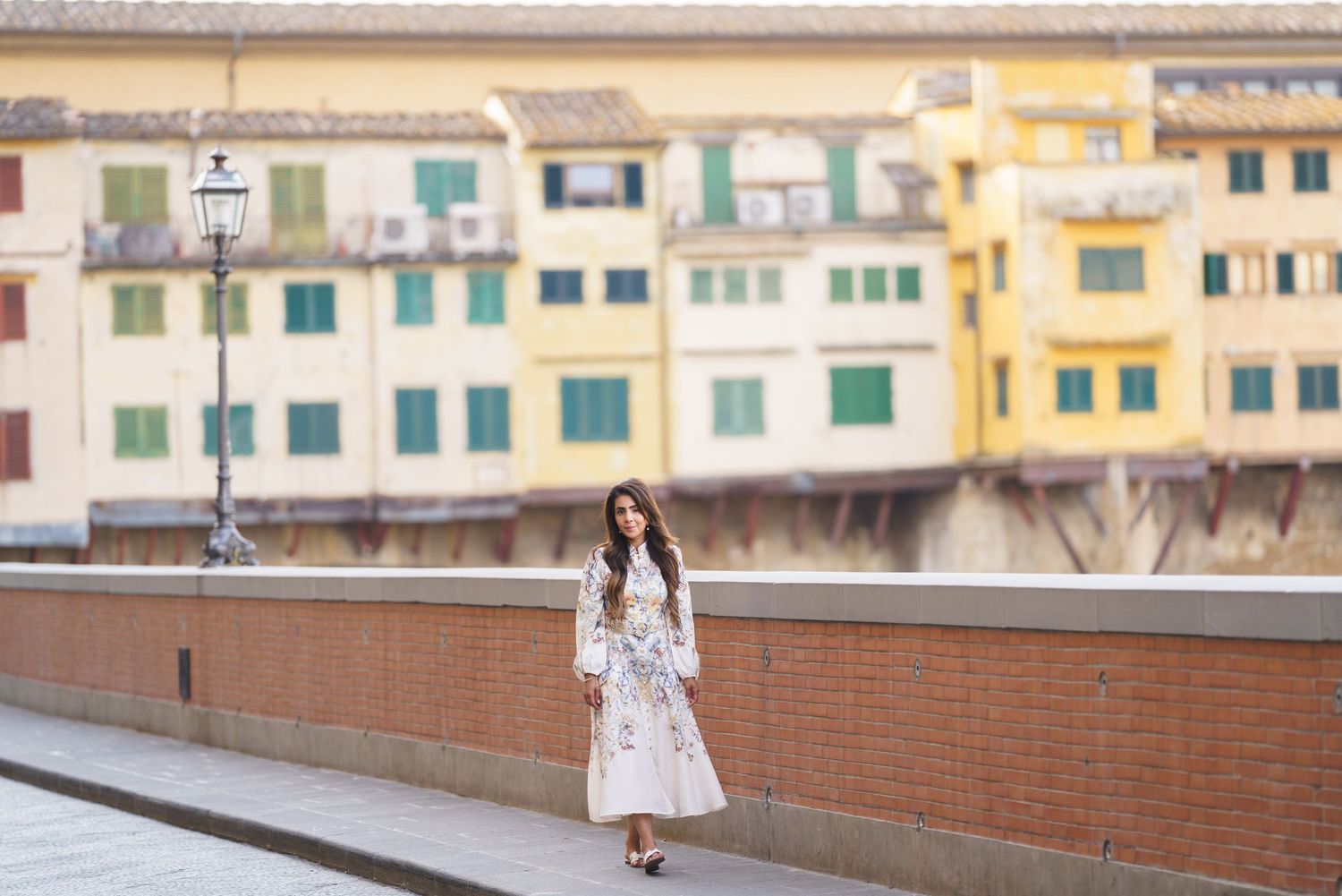 A woman in a white floral dress stands on a bridge in Florence, Italy, with colorful buildings in the background.