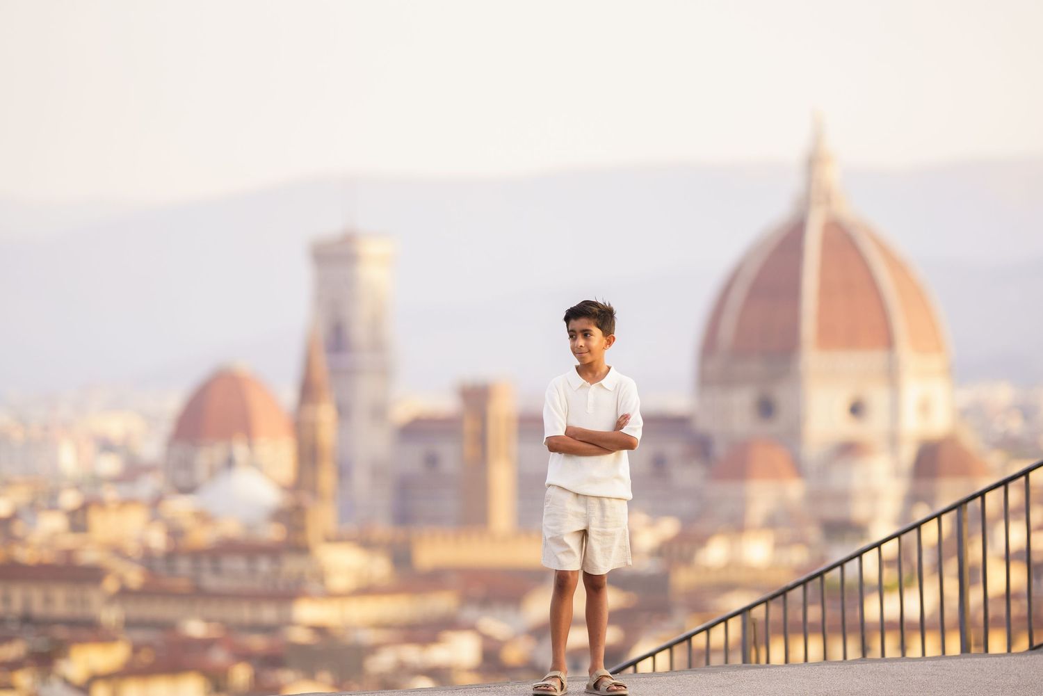 A young boy stands on a terrace with arms crossed, overlooking the Florence skyline with the iconic Duomo cathedral in the background.