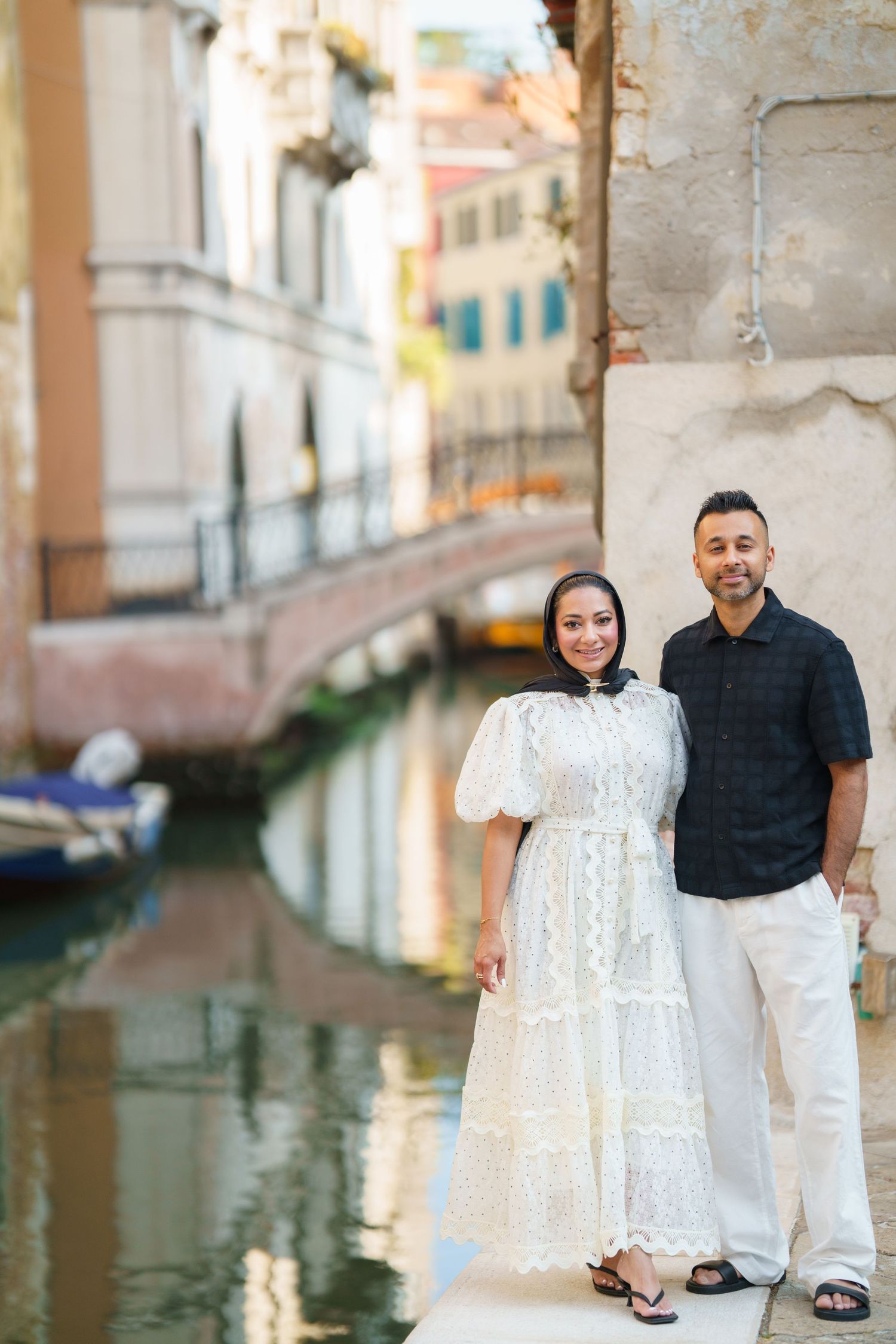 Couple posing by a Venice canal, woman in white lace dress, man in black shirt, colorful buildings reflected in water.
