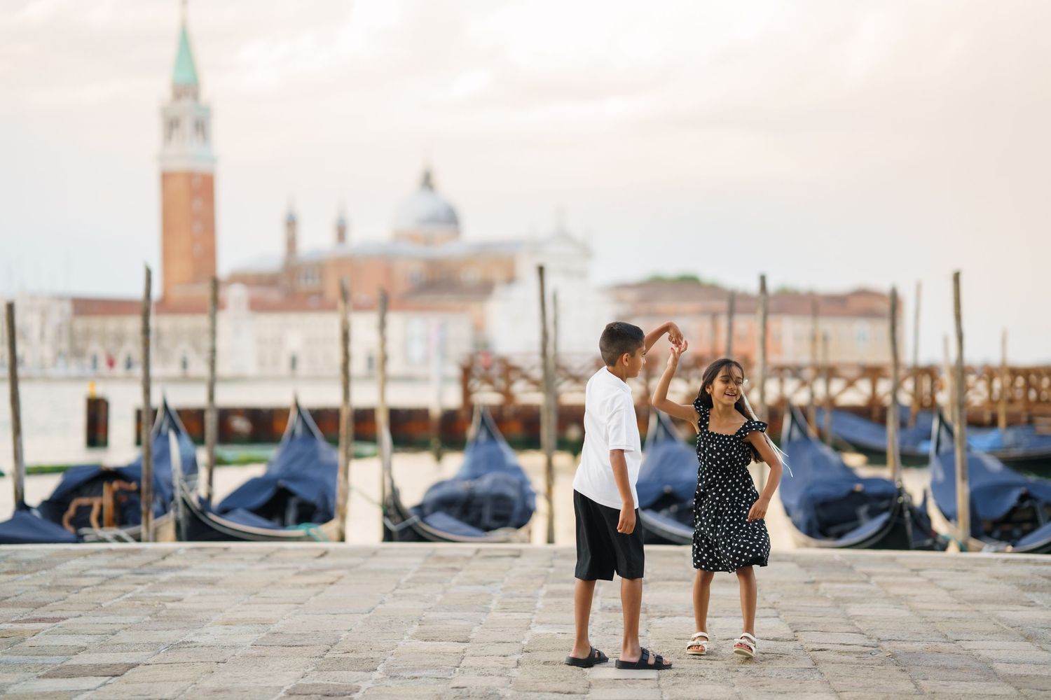 Two children pose on a Venice waterfront with gondolas and San Giorgio Maggiore church in the background.