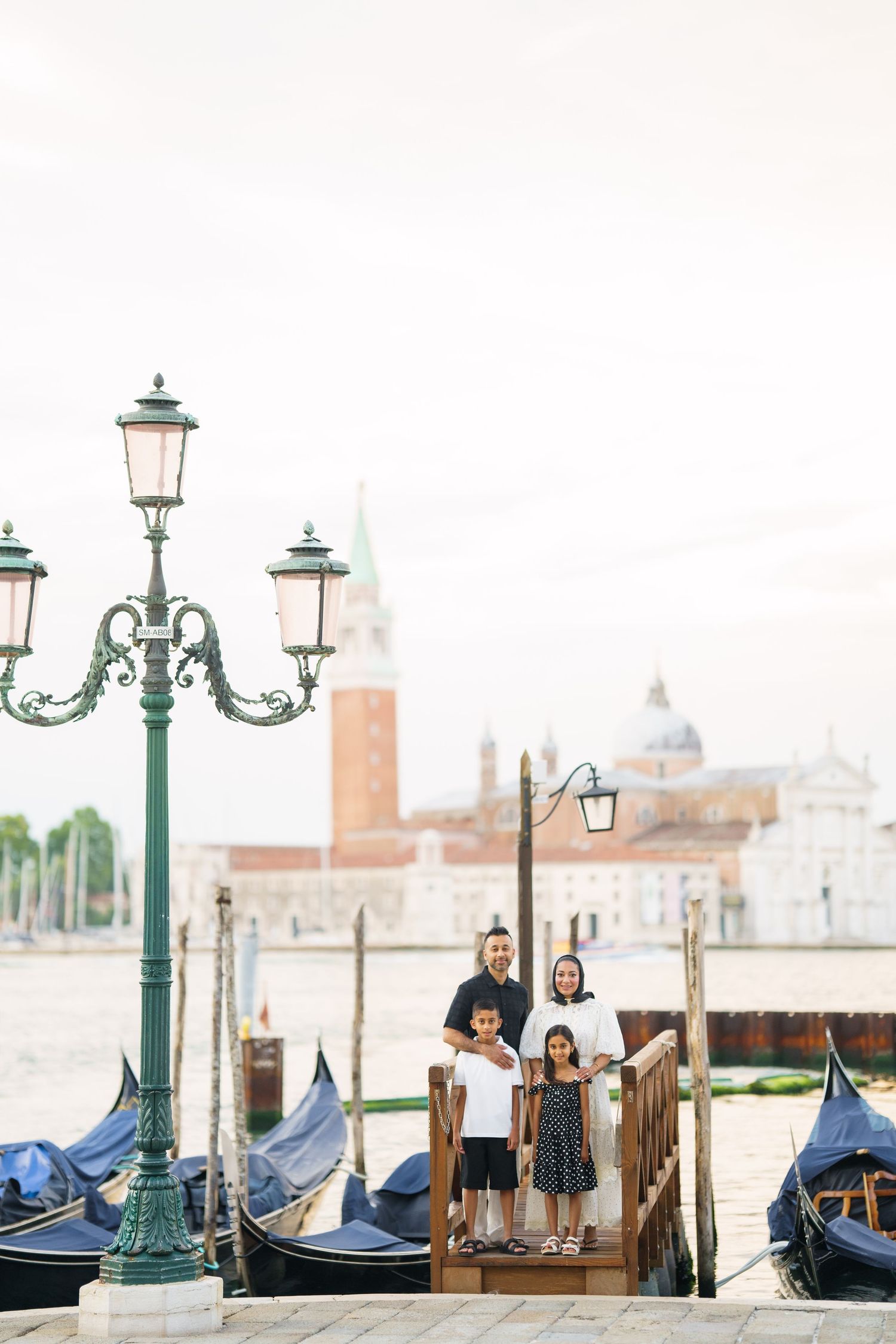 Family of four posing on a Venice dock with gondolas and San Giorgio Maggiore church in the background.