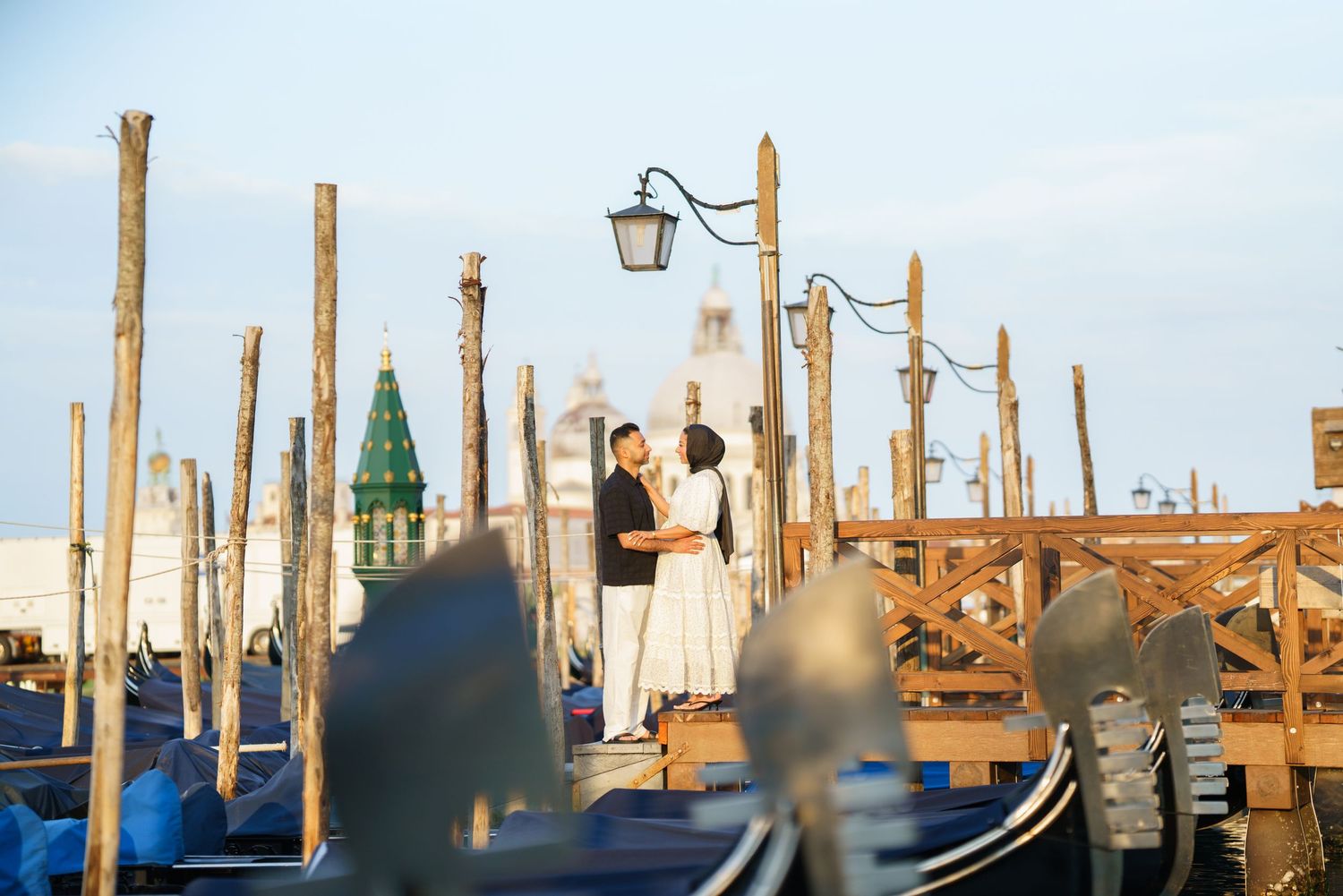 Couple embracing romantically among gondolas and wooden poles at Venice waterfront with historic architecture backdrop.