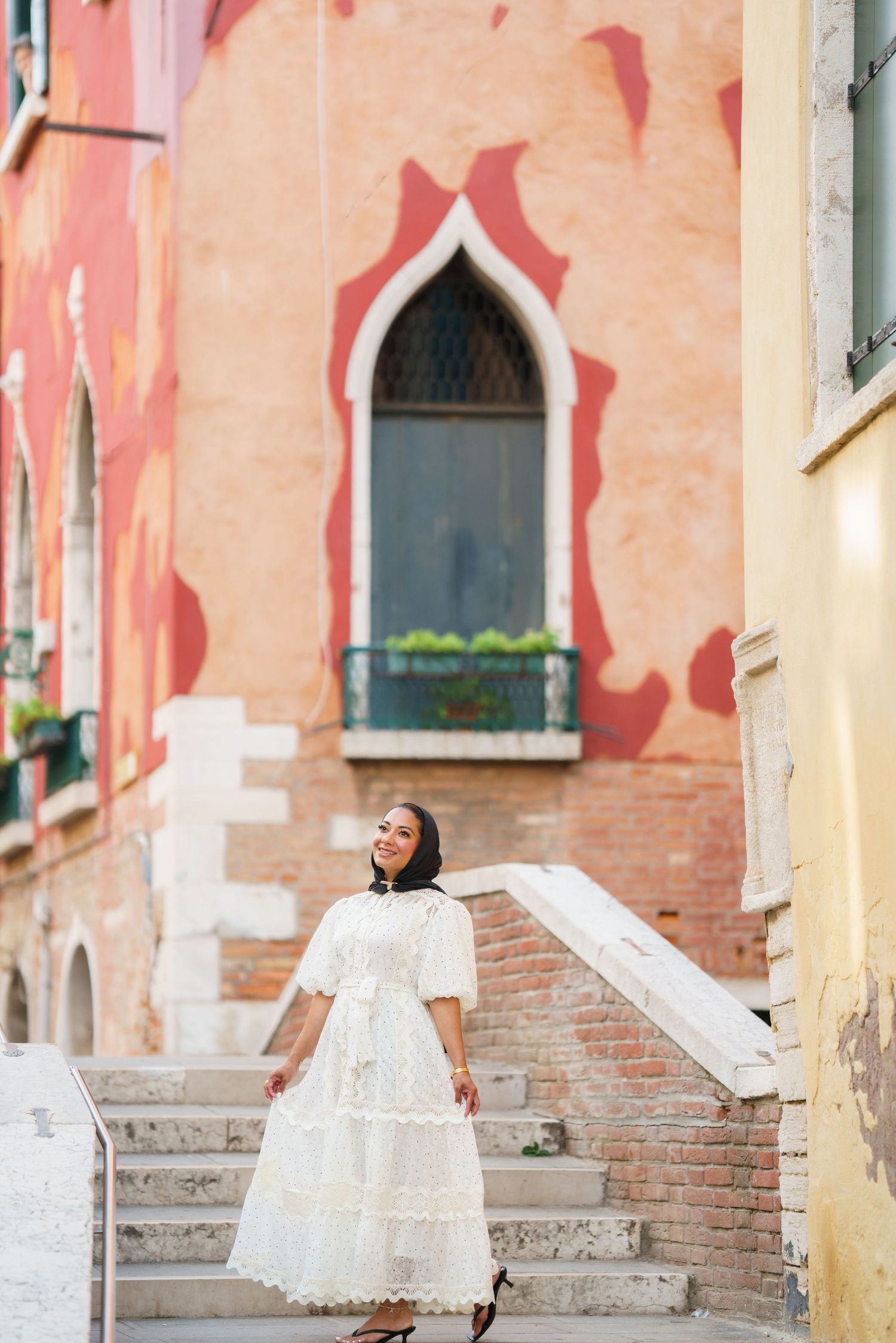 Woman in white lace dress smiling on stone steps before colorful Venetian building with Gothic window.