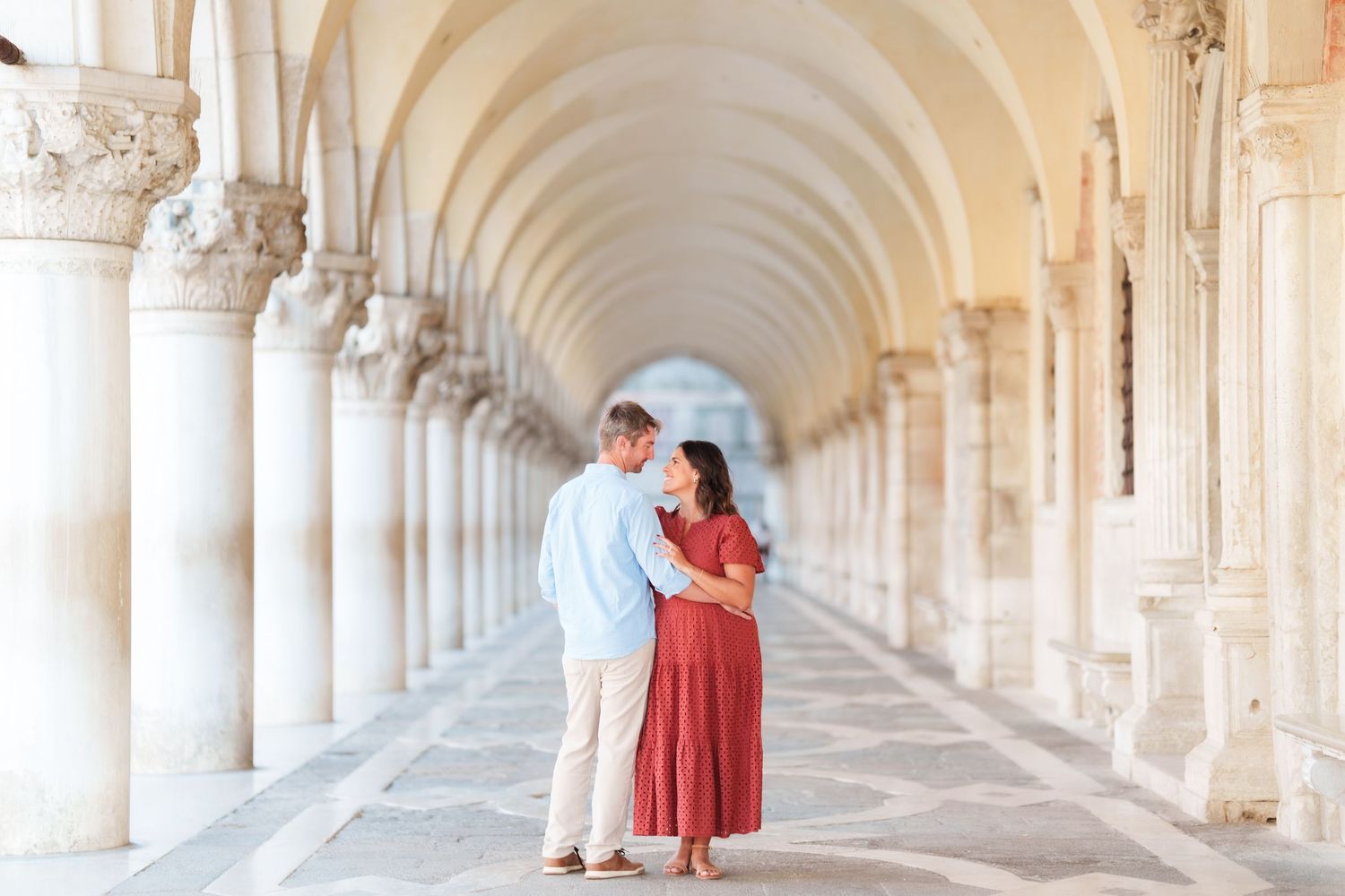 Couple embracing under elegant arched colonnade in Venice, Italy, woman in red dress and man in light blue shirt.