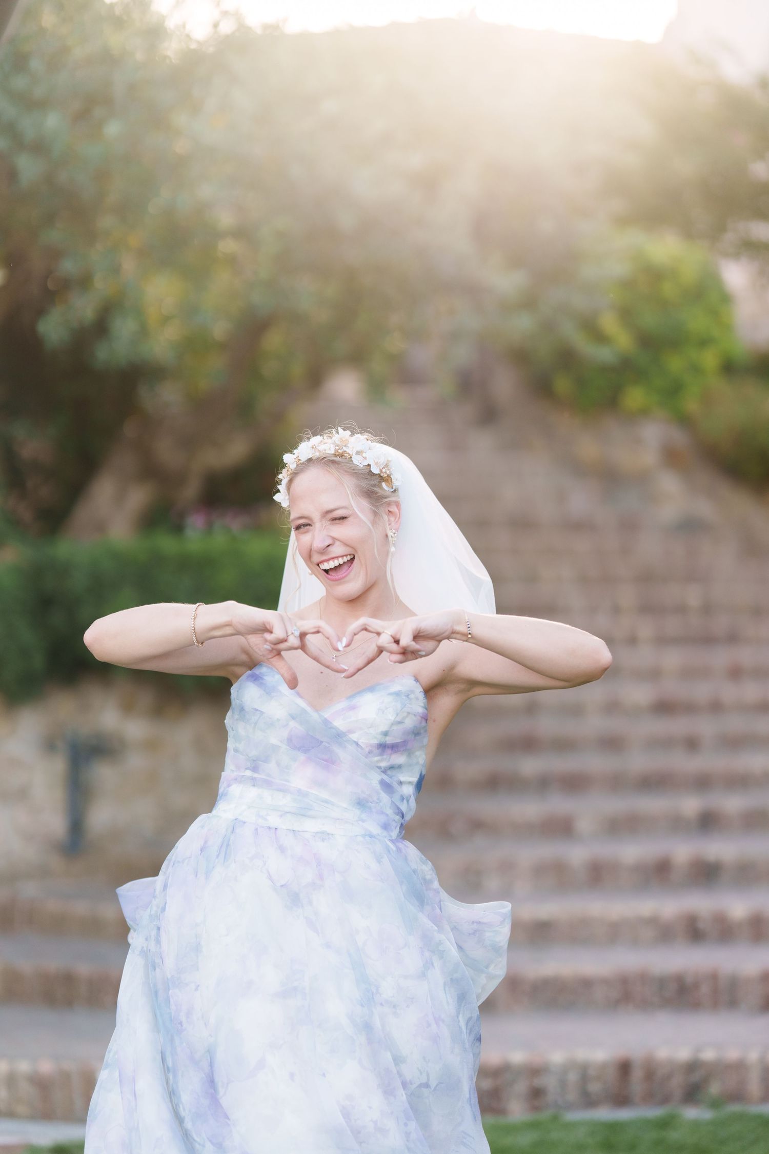 Smiling bride in floral white dress and veil makes heart shape with hands outdoors on sunny day.