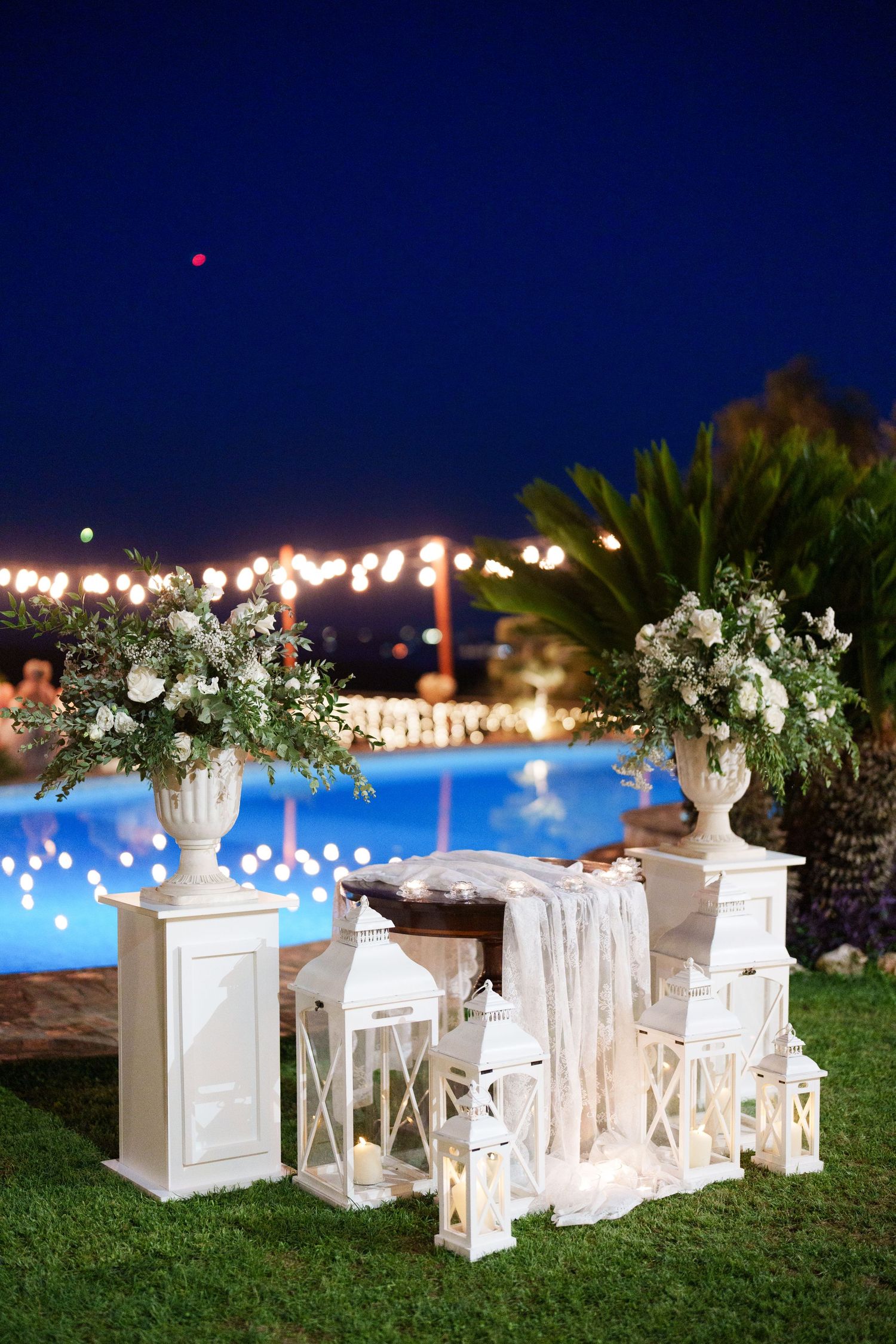Elegant outdoor wedding reception setup at night with white floral arrangements, lanterns, and string lights near a pool.