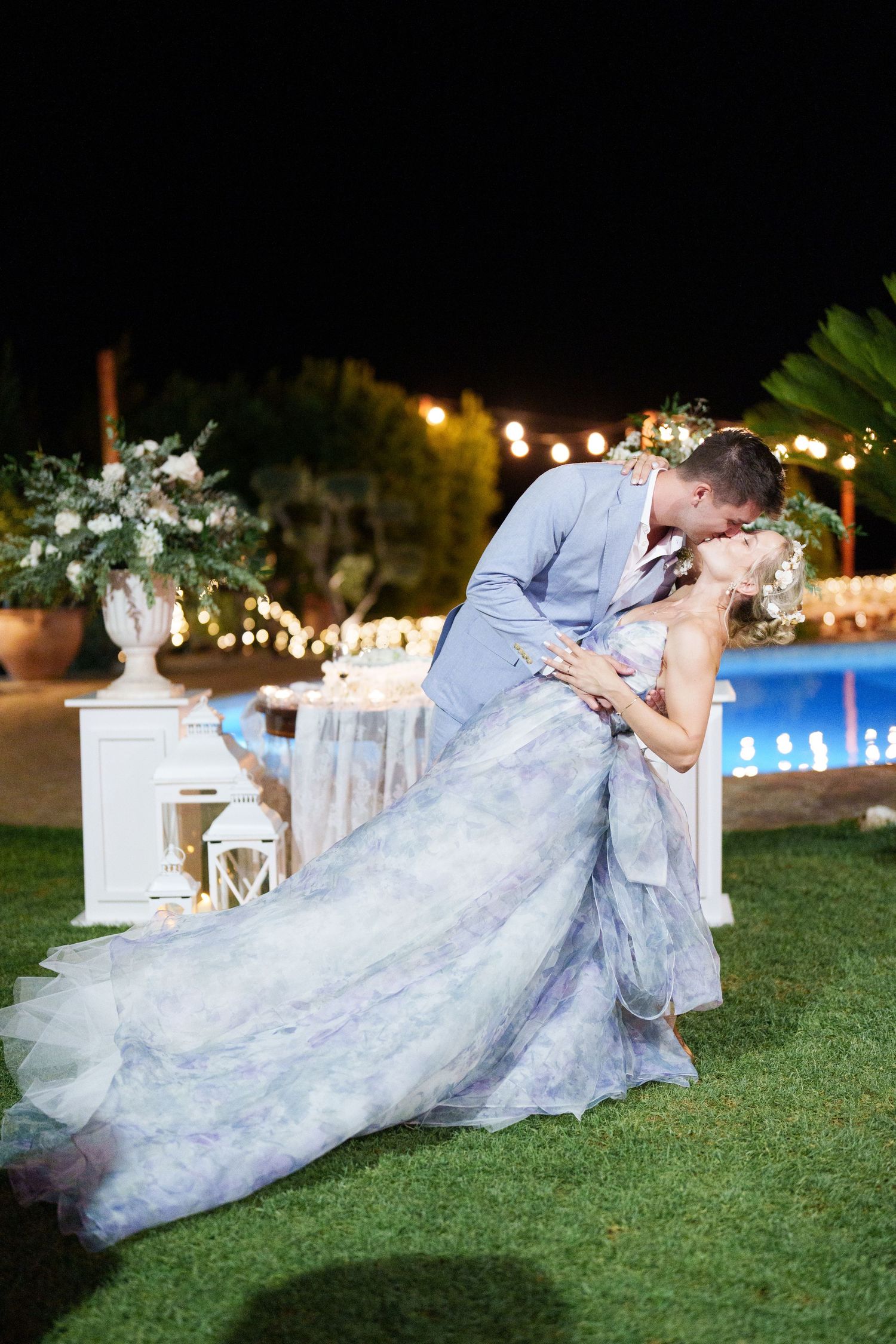 Bride and groom share a romantic dip kiss at night, with pool and string lights glowing behind them.